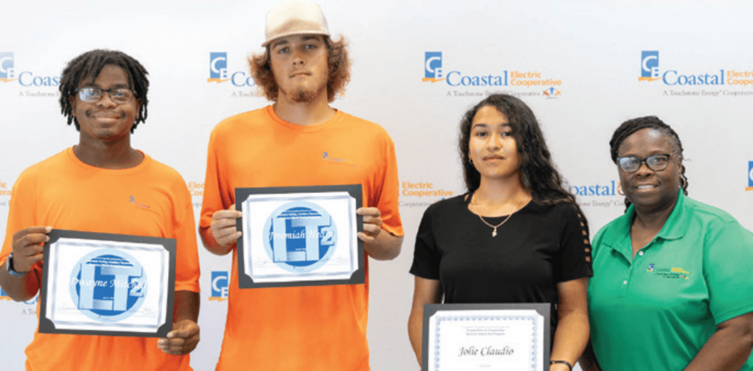 Four people stand in front of a "Coastal Electric Cooperative" backdrop, holding certificates, with the two center people wearing orange shirts.