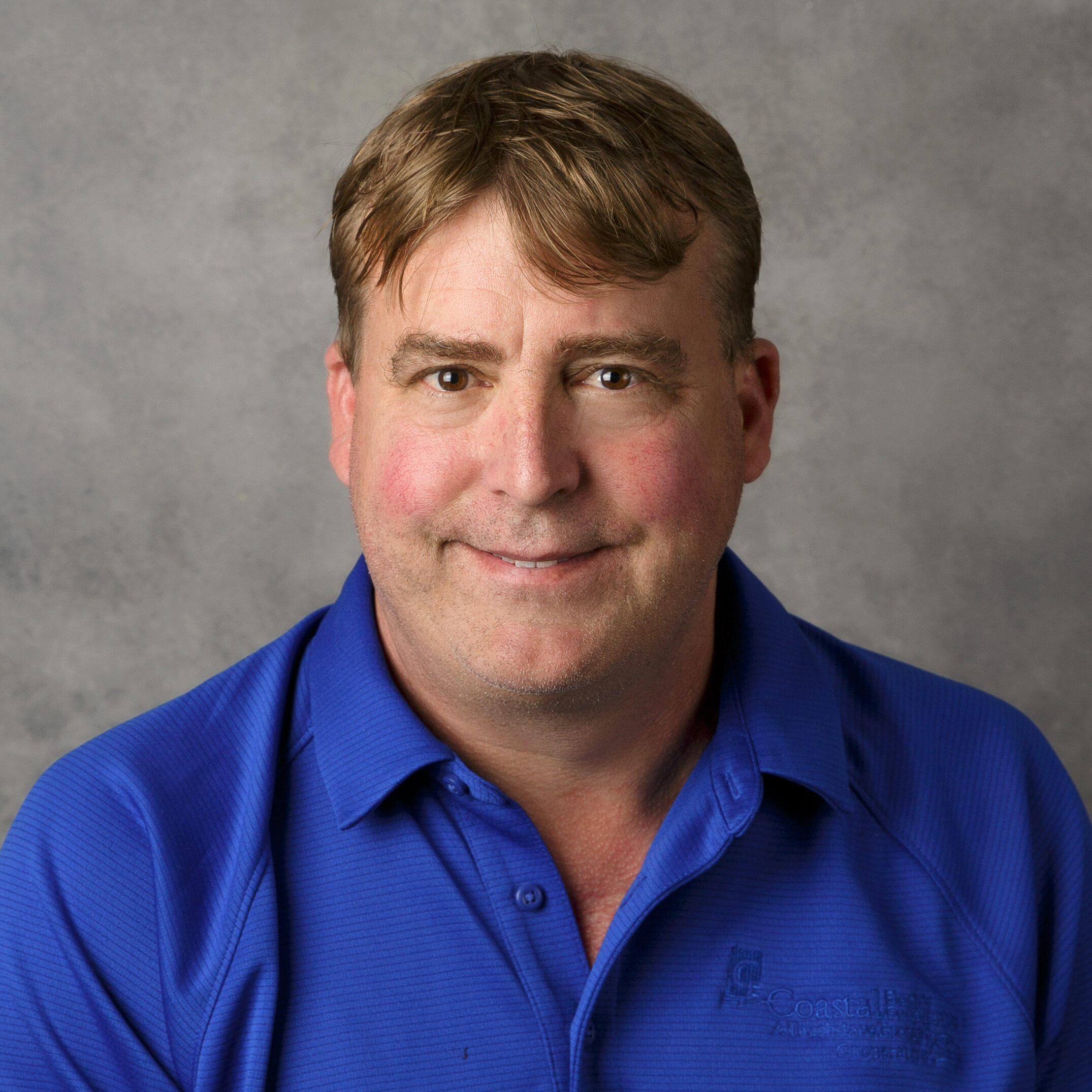 A person in a blue shirt against a gray background smiles subtly. The shirt has a logo on the left side.