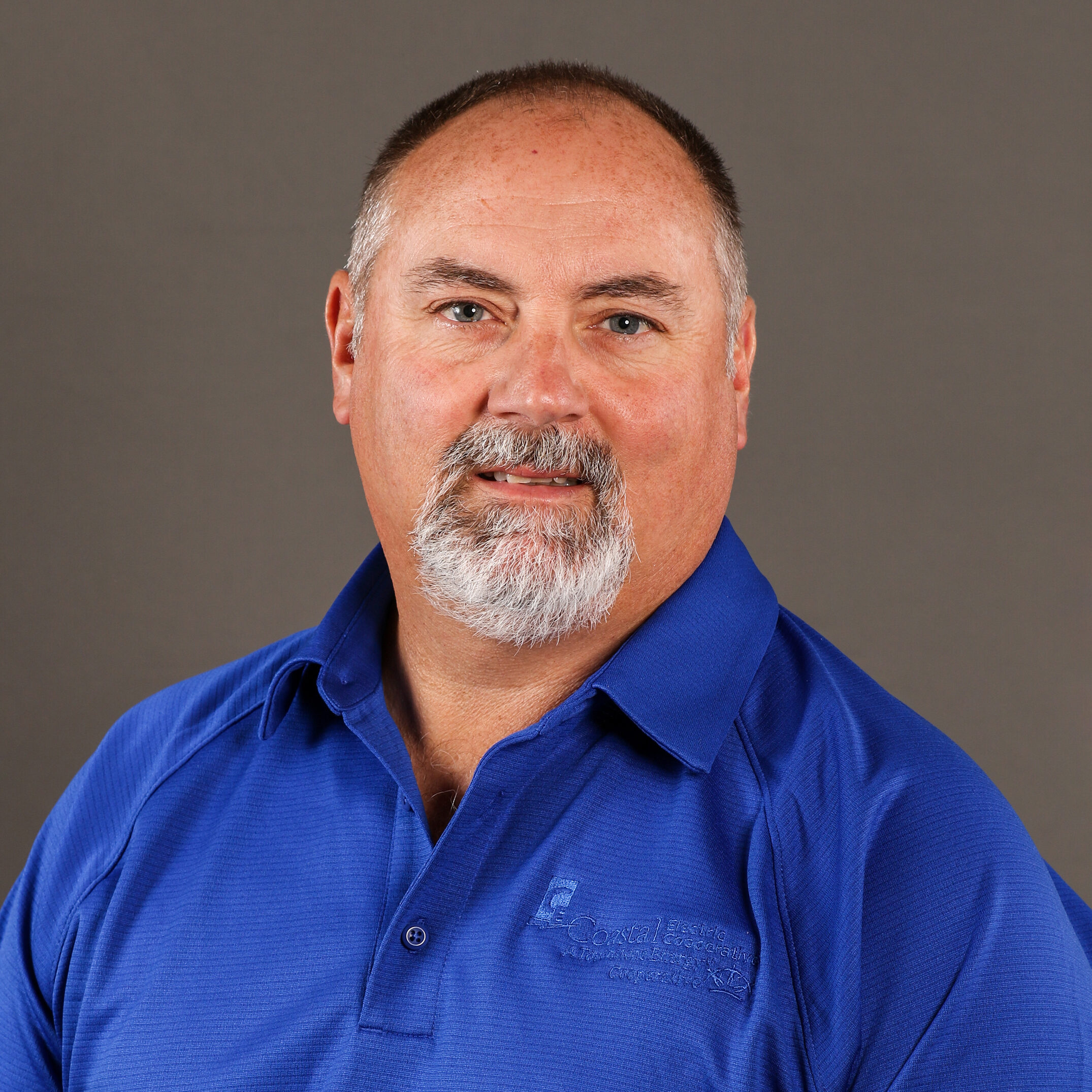 A person with a goatee wears a blue polo shirt with a logo on a plain grey background. No landmarks or buildings visible.