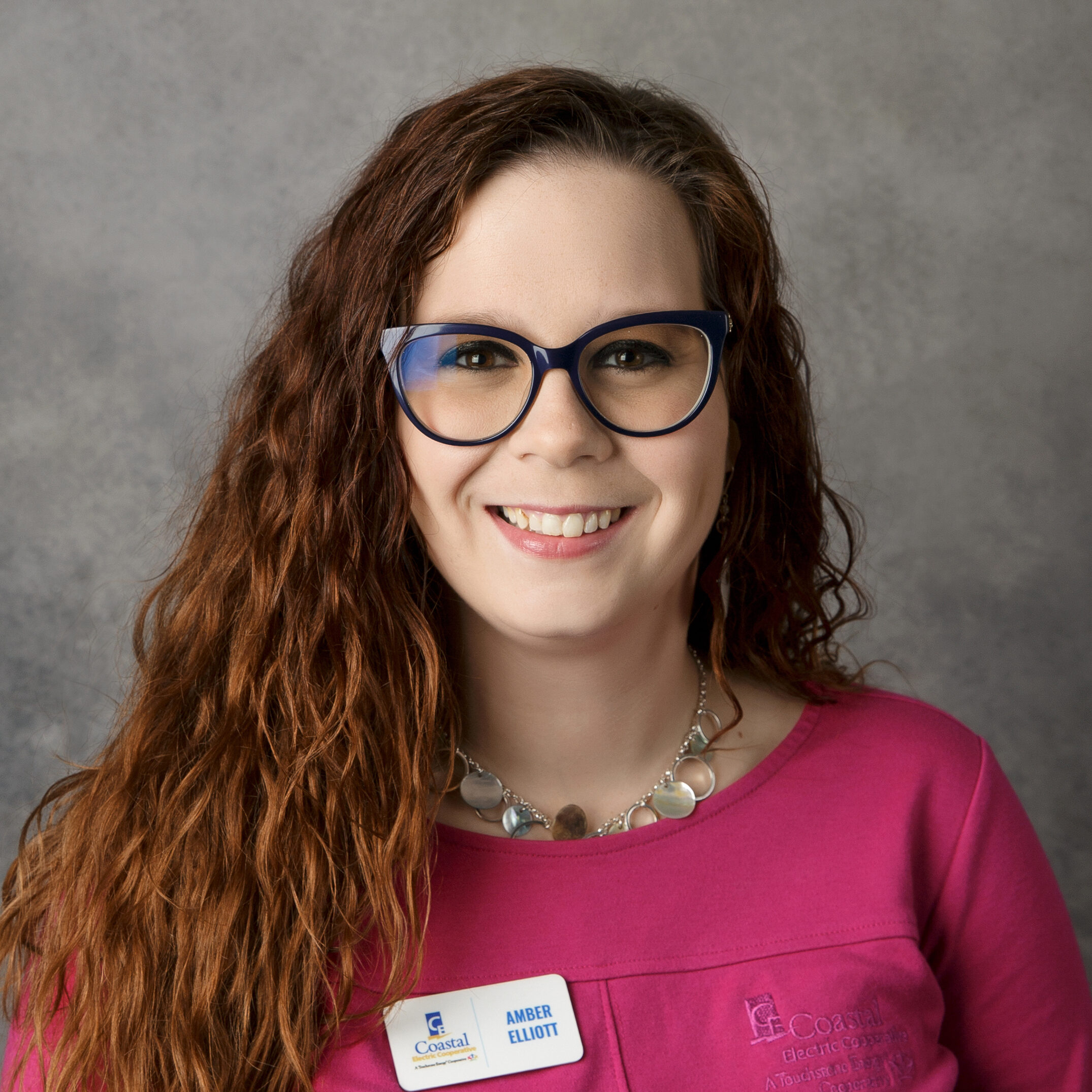 A person with long, wavy hair and glasses smiles at the camera, wearing a pink top and a name tag reading "Amber Elliott."