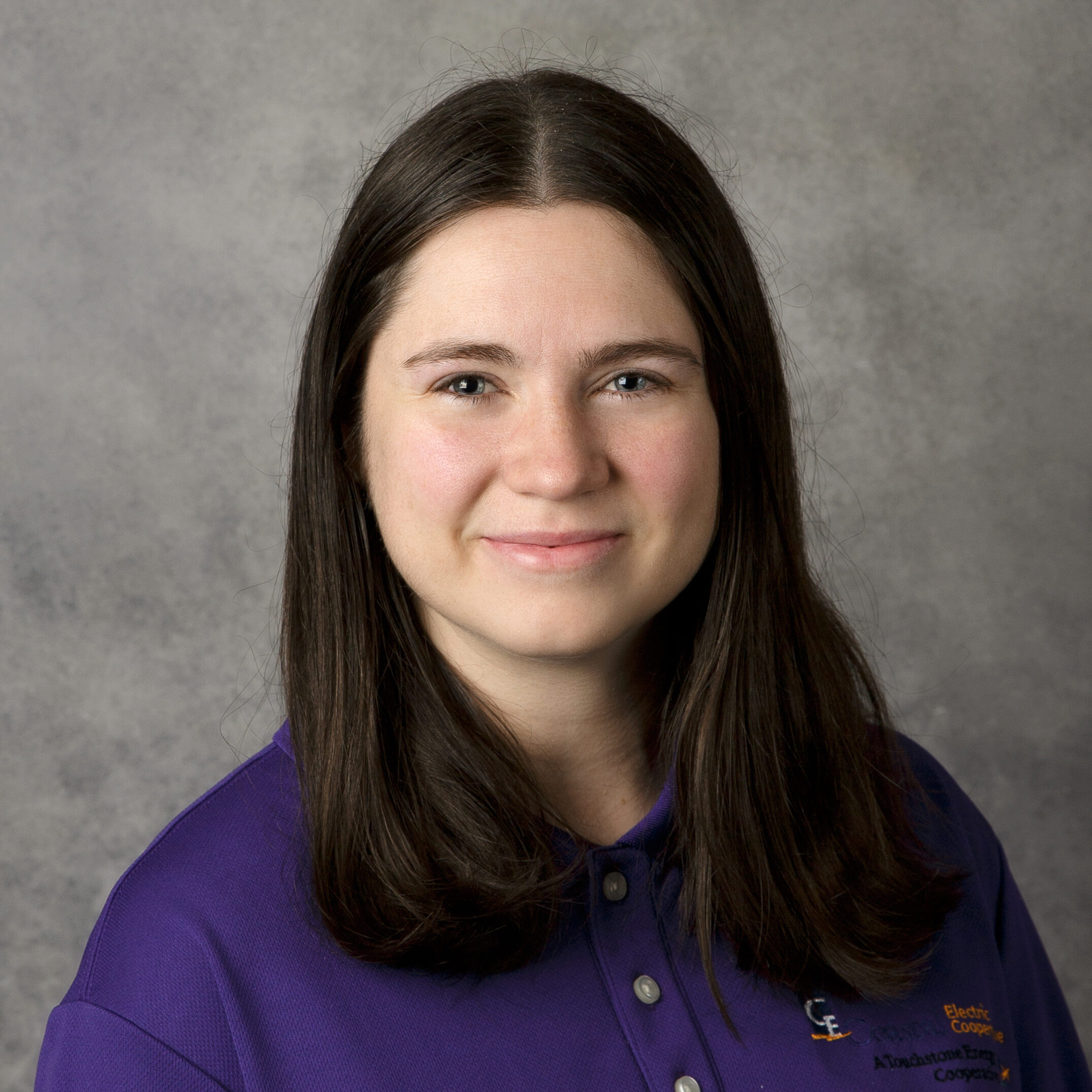 A person with long dark hair wearing a purple shirt against a grey background, smiling slightly at the camera.