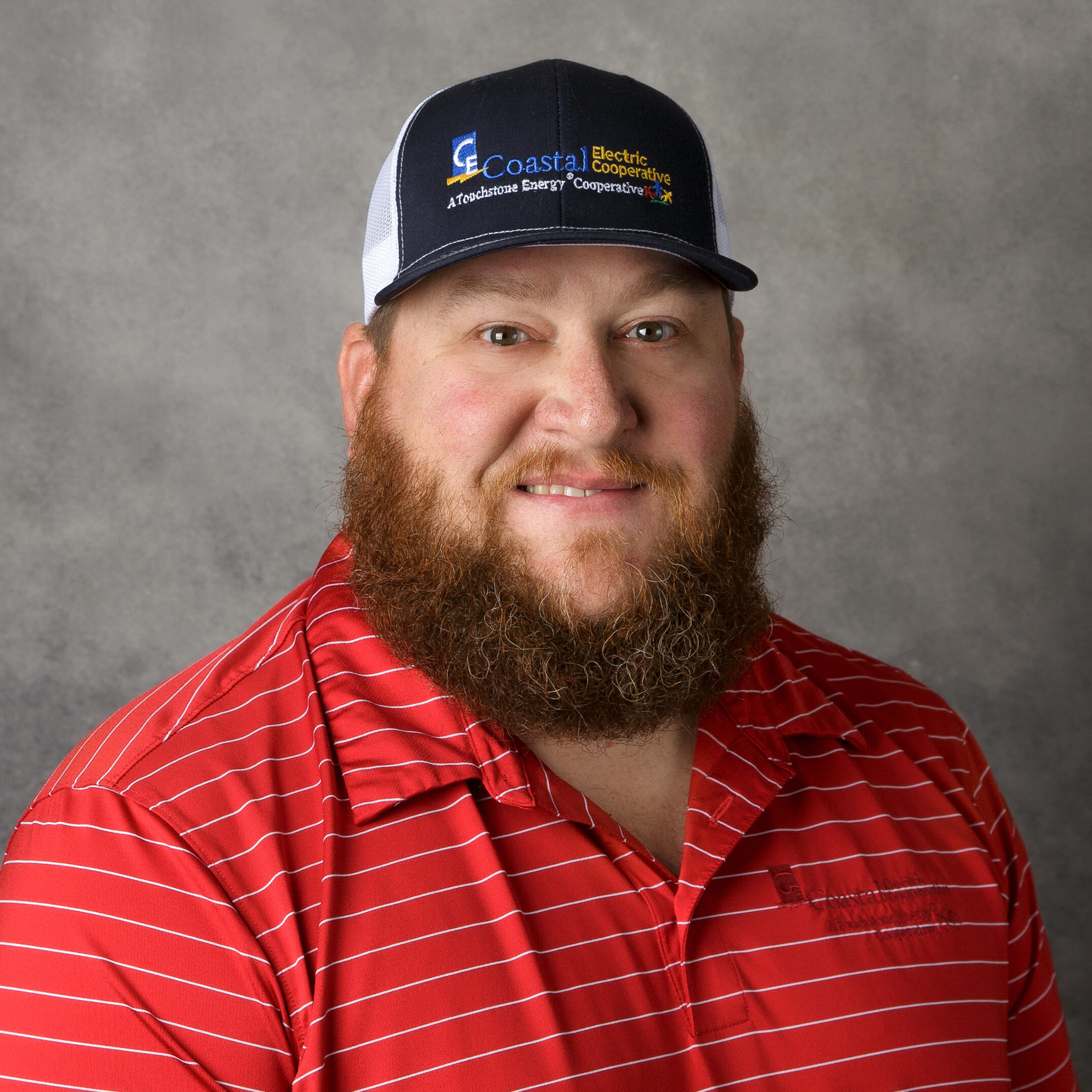 A person with a beard wears a Coastal Electric Cooperative cap and a red striped shirt, smiling against a neutral gray background.