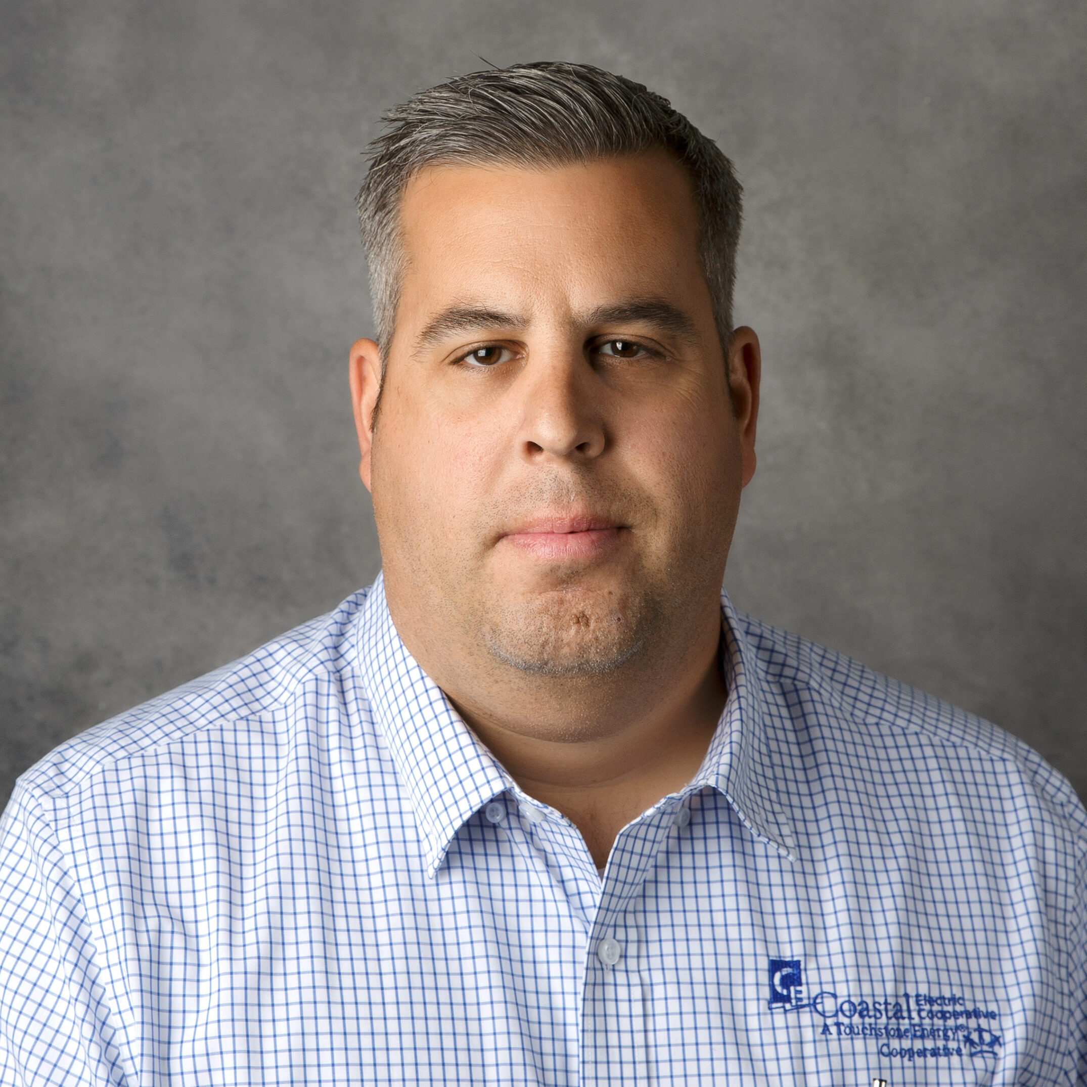 A person with short gray hair wears a checkered shirt with a logo, set against a plain gray background.