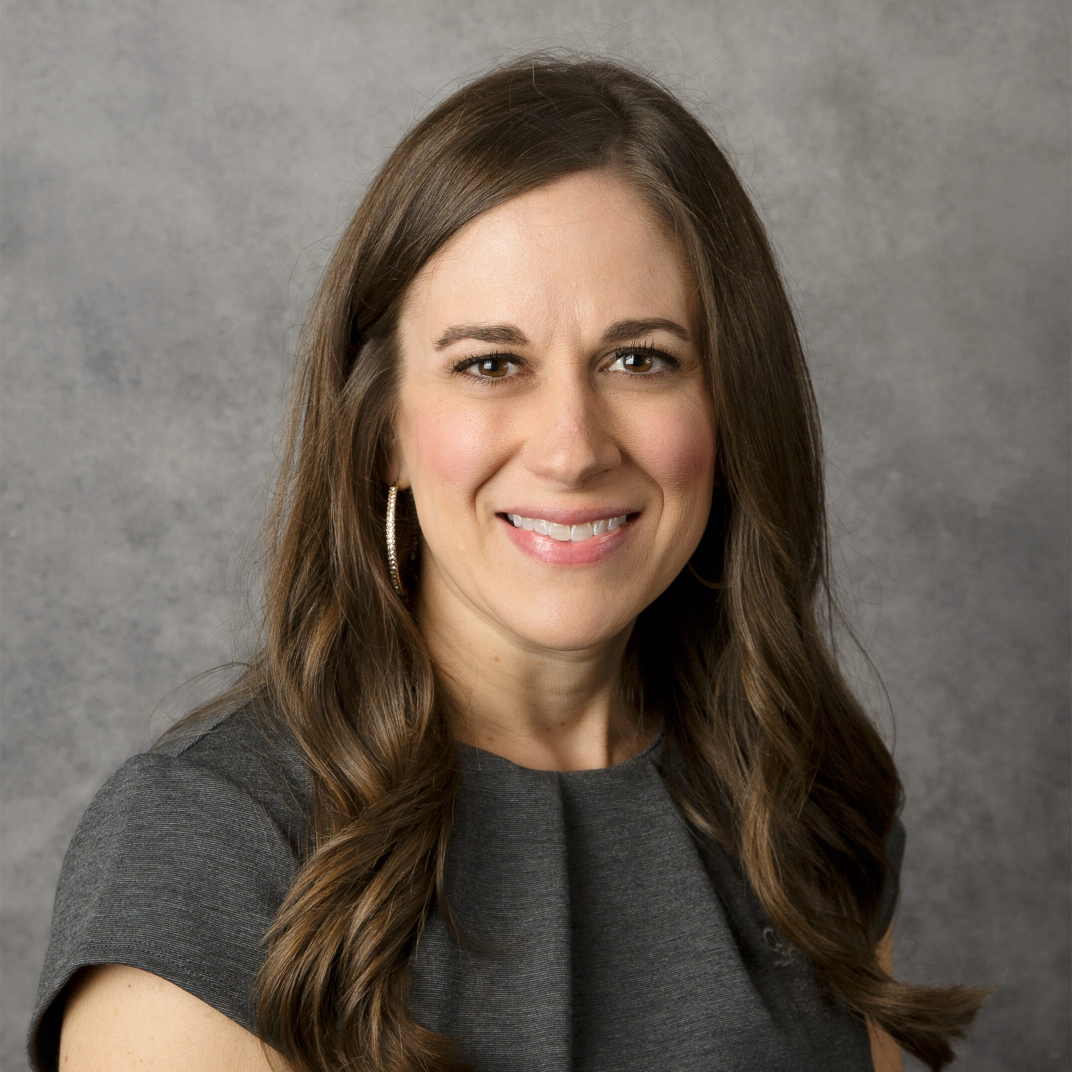 A person with long brown hair and hoop earrings smiles against a neutral gray background, wearing a dark gray top.