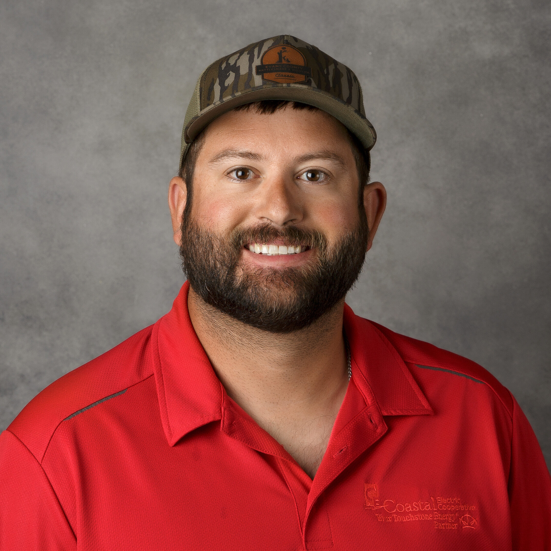 A person wearing a camo hat and red shirt is smiling against a plain gray background. The shirt has a logo on it.