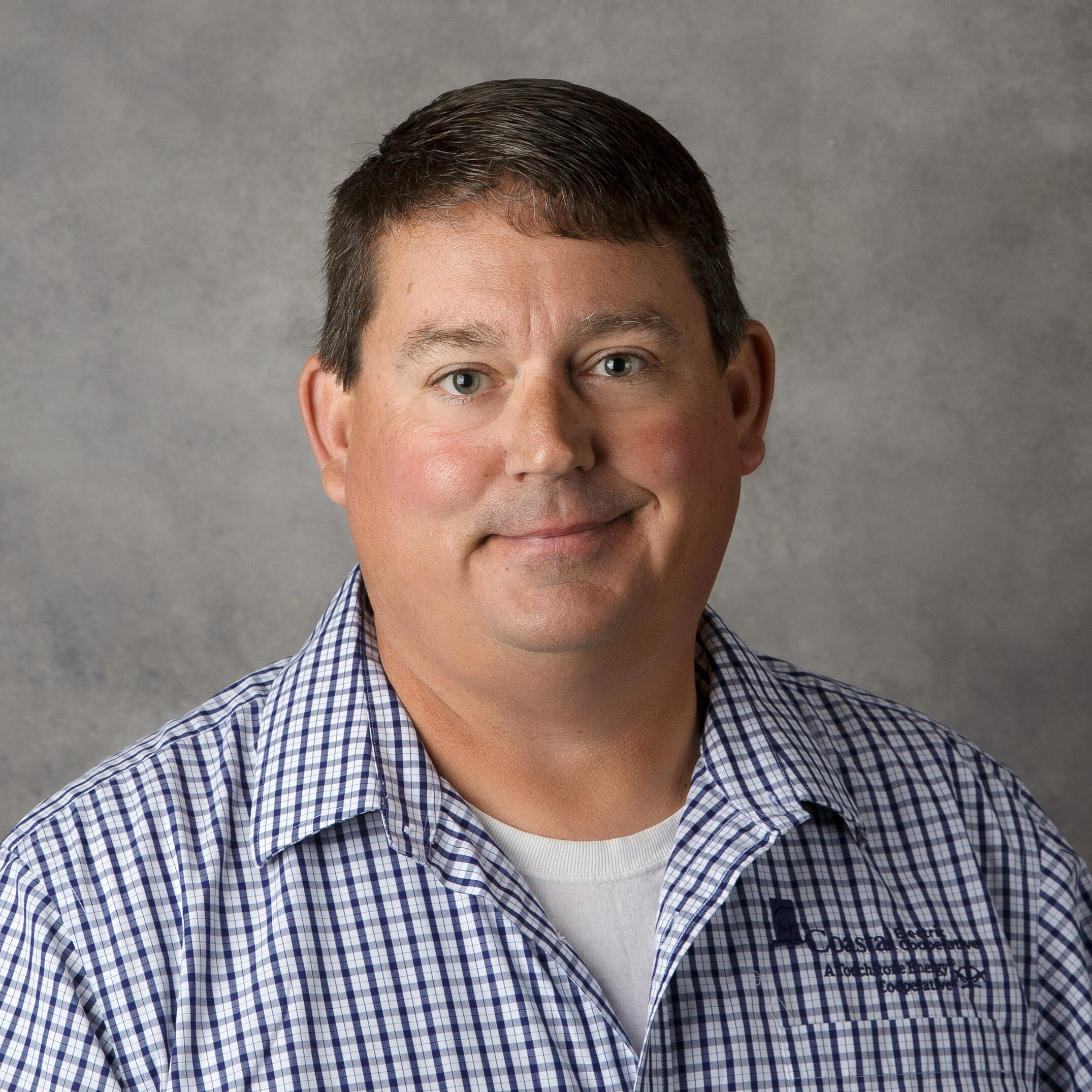 A person with short hair and a checked shirt stands against a plain background, smiling slightly, with no visible landmarks or buildings.