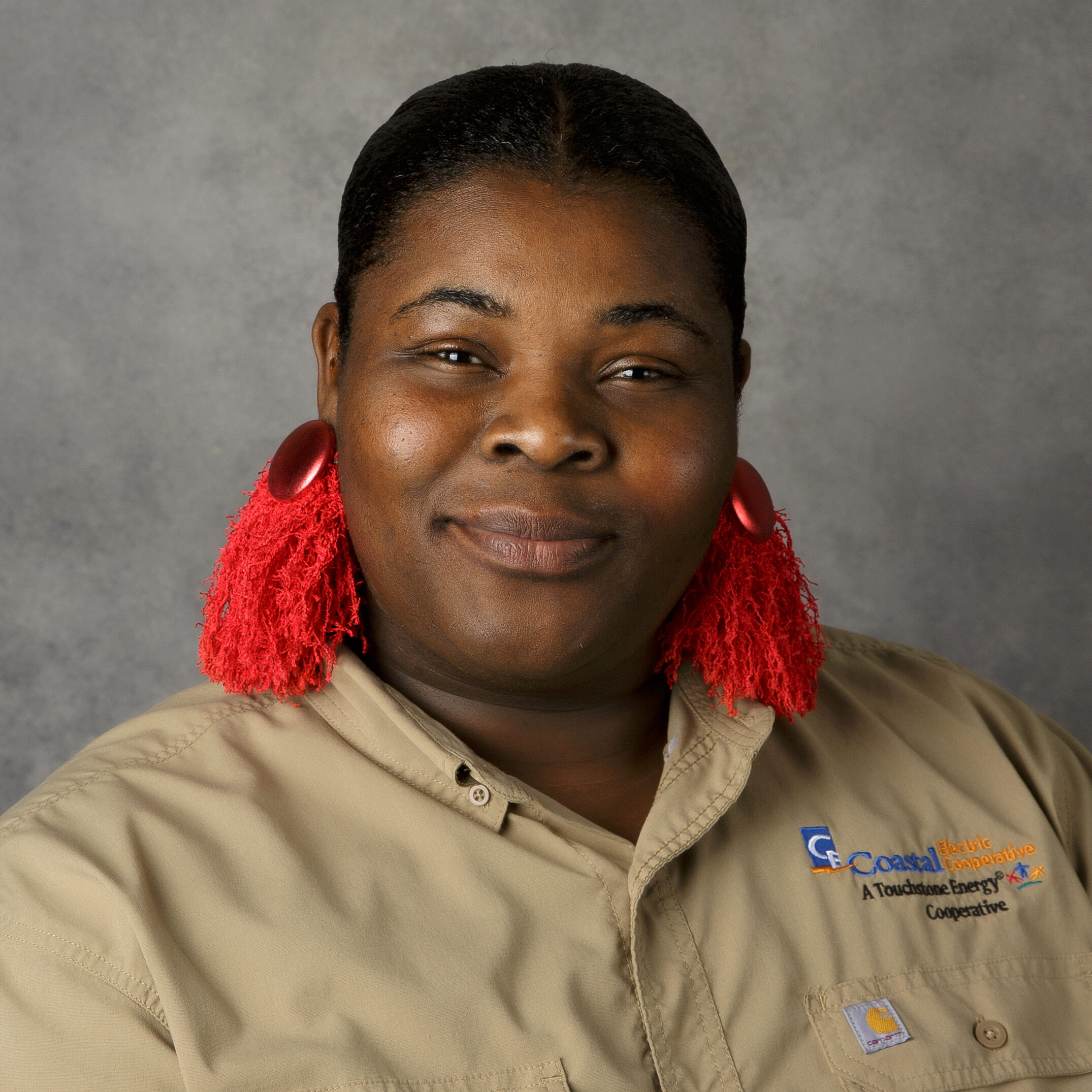 A person with red earrings wearing a beige Coastal Electric Cooperative shirt, smiling against a gray background.