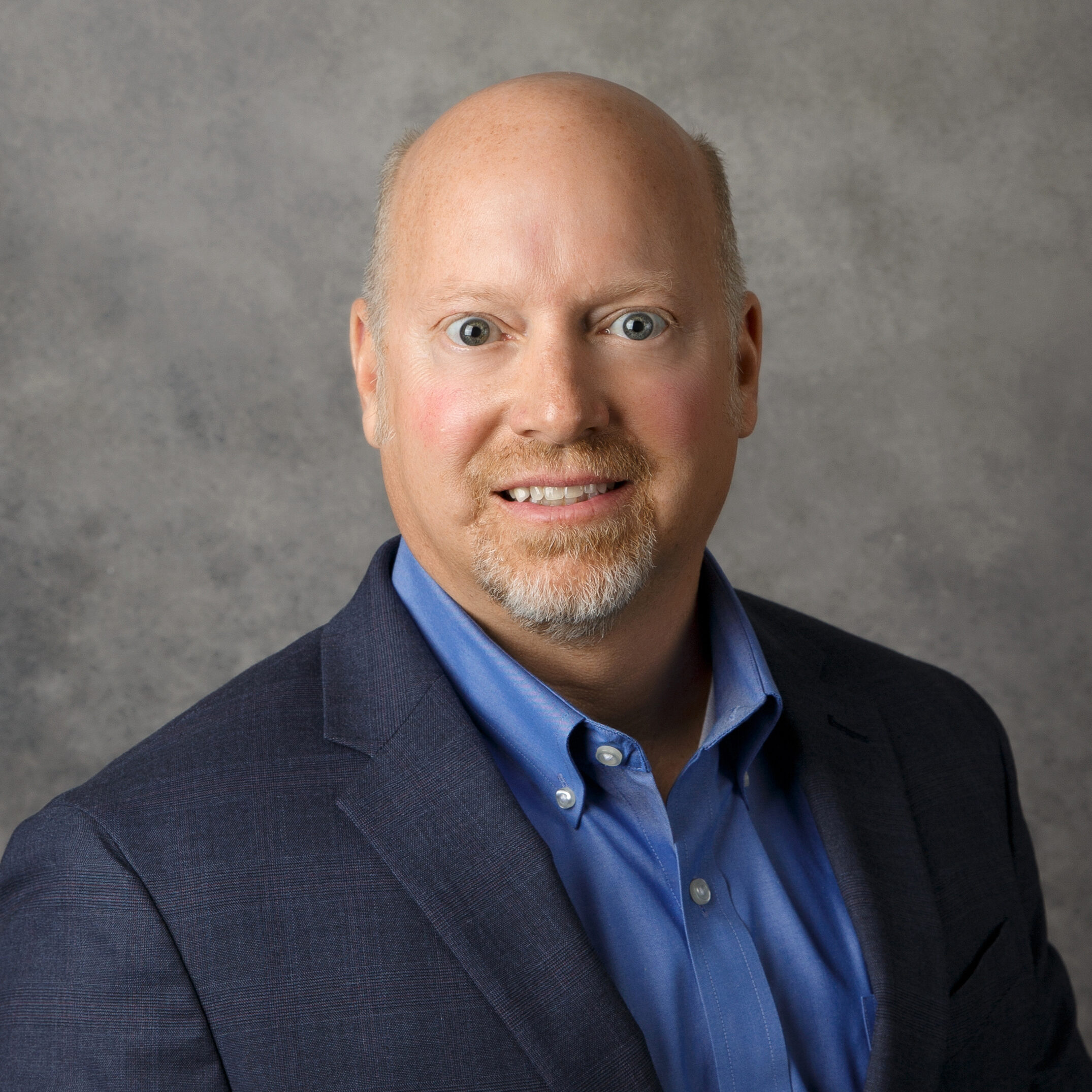 A person with a light beard and blue shirt is wearing a dark suit jacket against a plain gray background, looking directly at the camera.