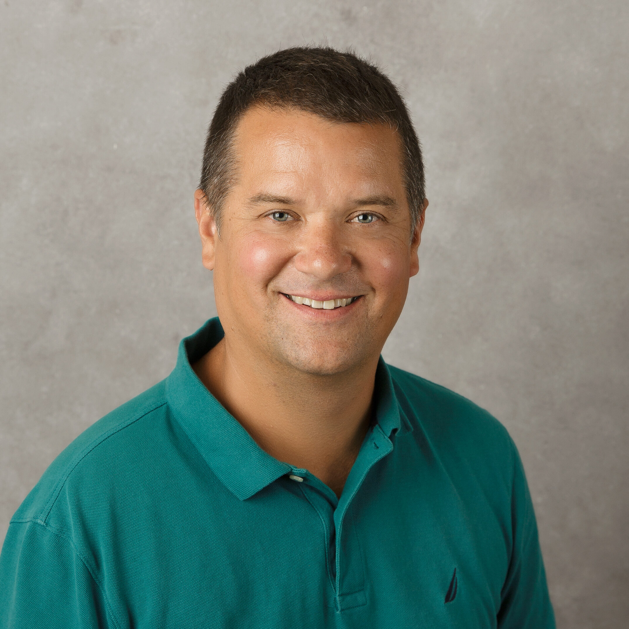 A person in a green shirt smiling against a plain, gray background. No recognizable landmarks or historical buildings are visible.