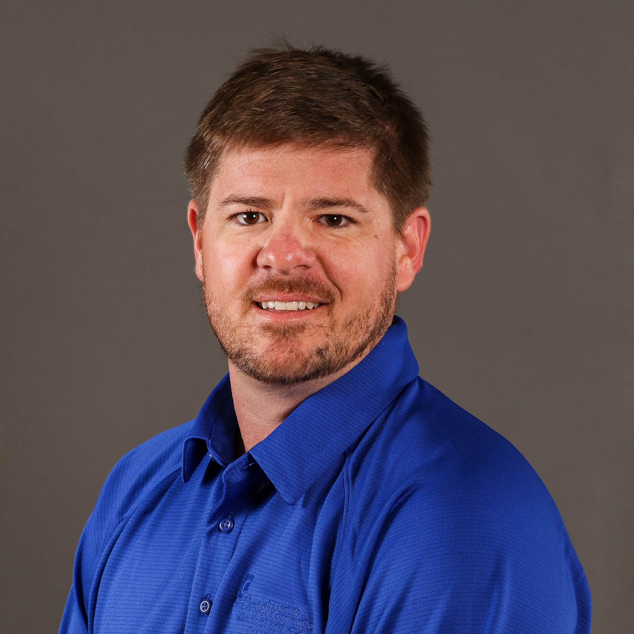 A person in a blue collared shirt stands against a plain gray background, looking toward the camera with a slight smile.