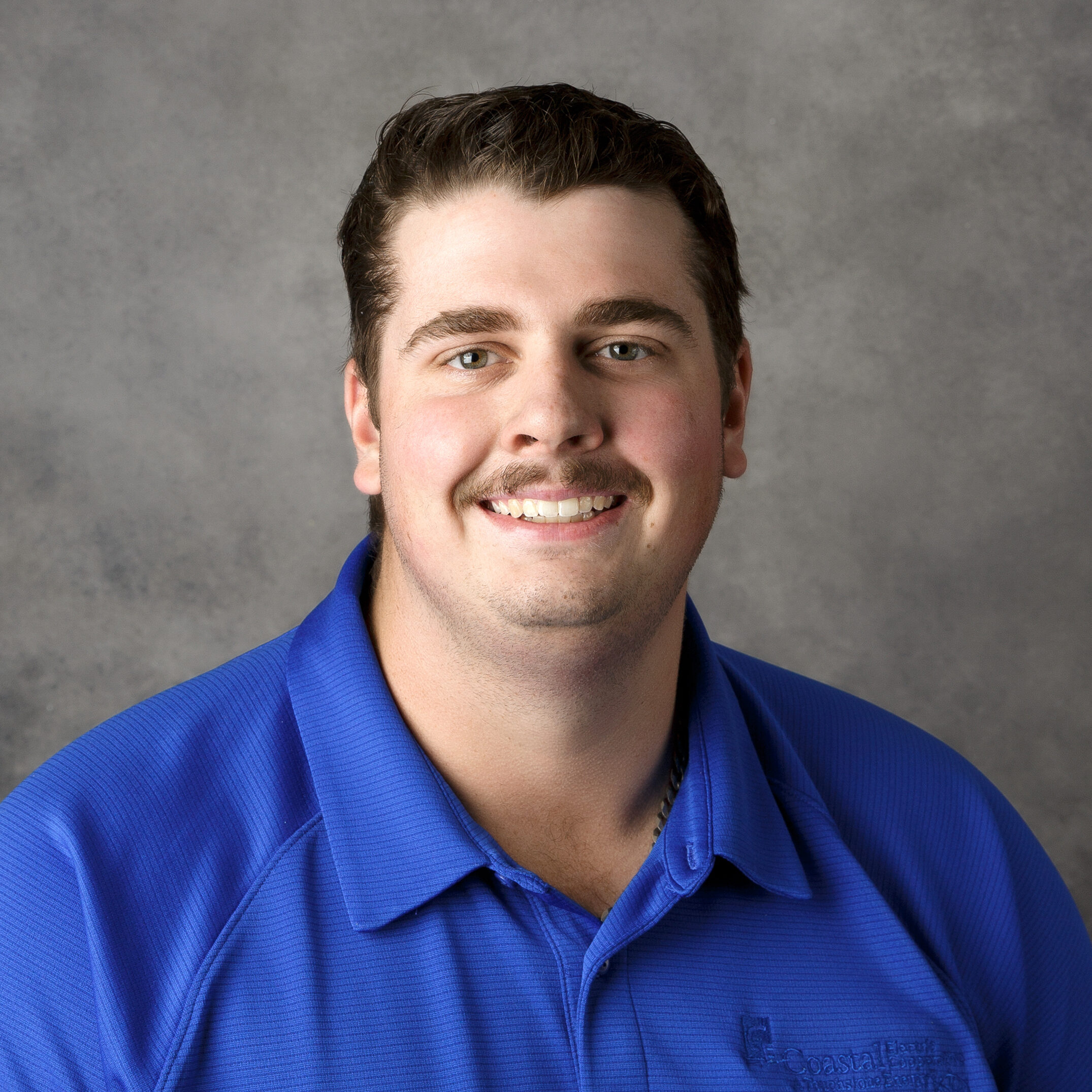 A person with a mustache smiles, wearing a blue polo shirt with a logo, against a gray background.