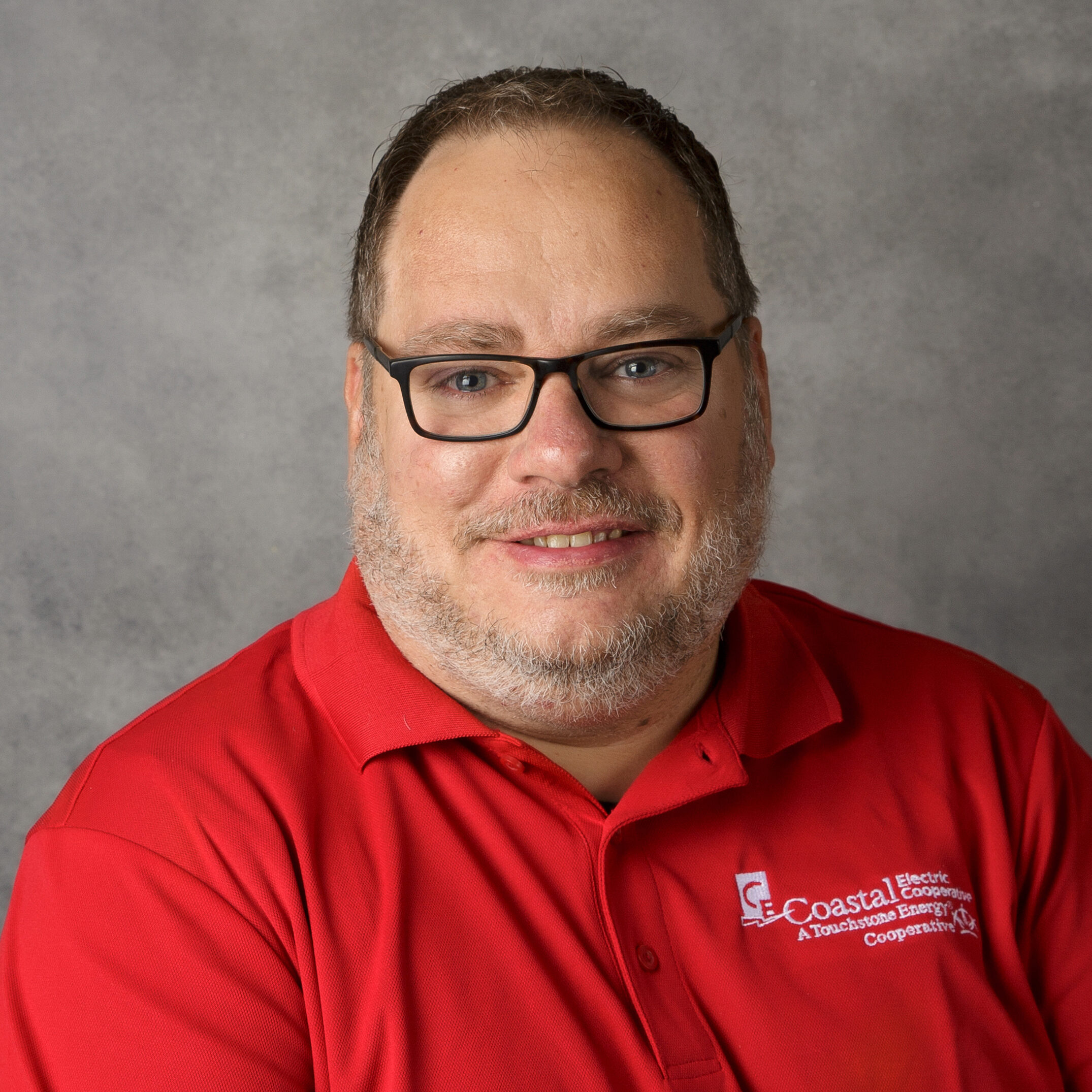 A person with glasses and a beard smiles, wearing a red "Coastal Electric Cooperative" shirt. Gray background provides a neutral backdrop.