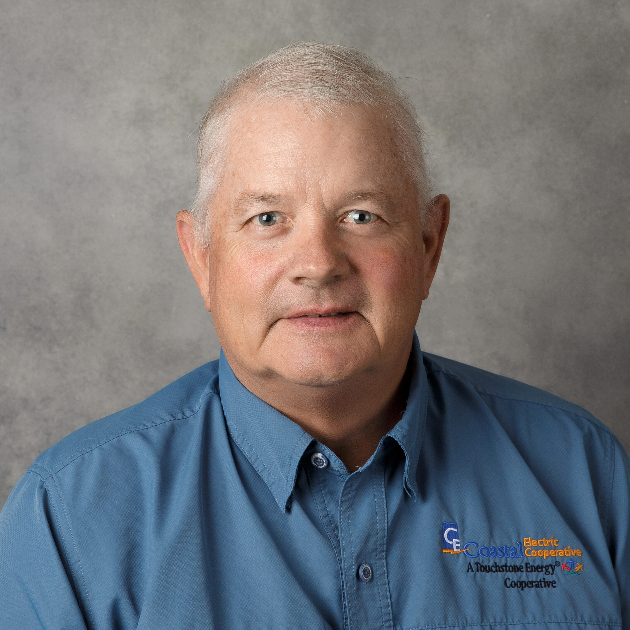 Person wearing a blue shirt with "Coastal Electric Cooperative" logo, seated against a neutral gray background, looking towards the camera.