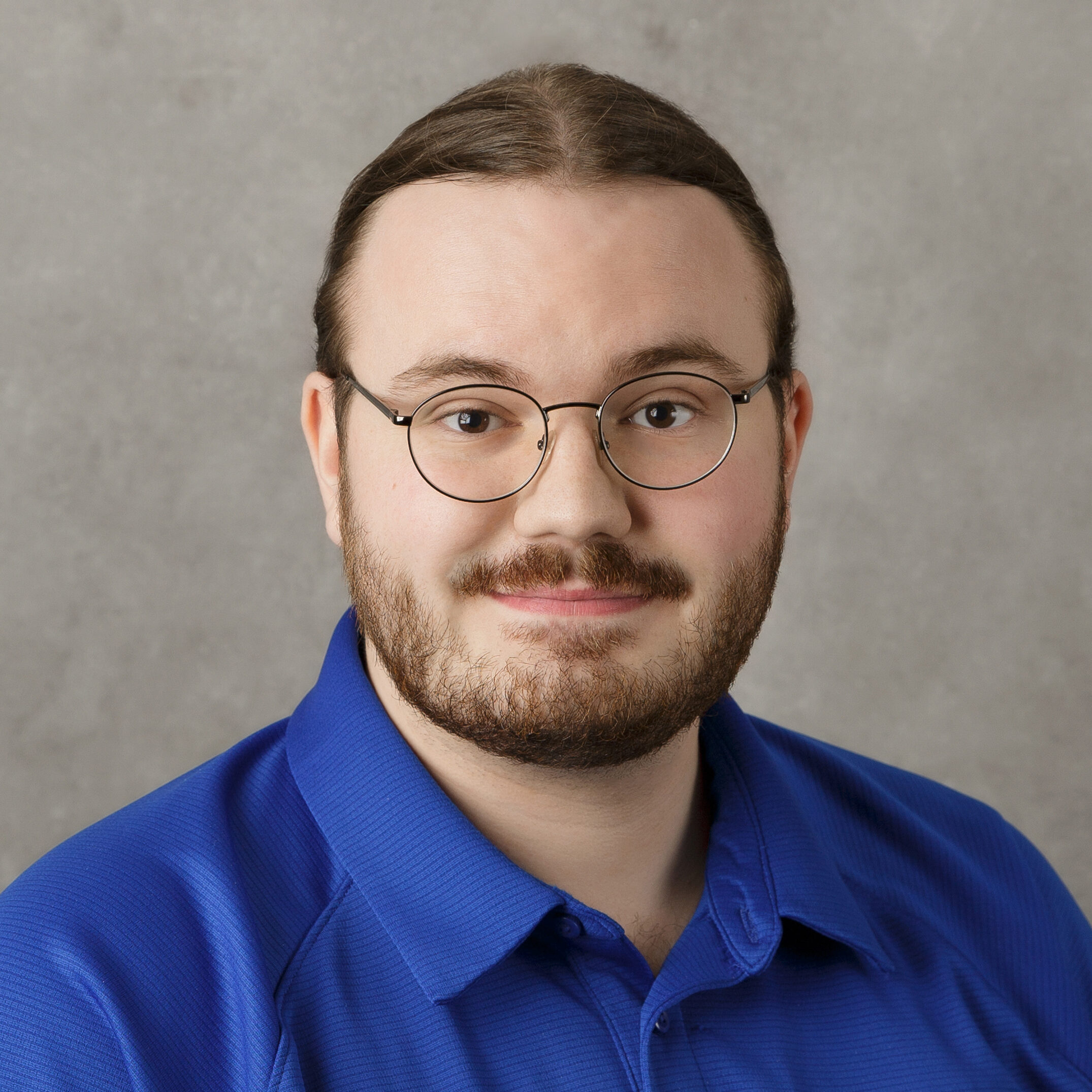 A person with glasses and a beard, wearing a blue shirt, stands against a plain gray background. No landmarks or buildings visible.