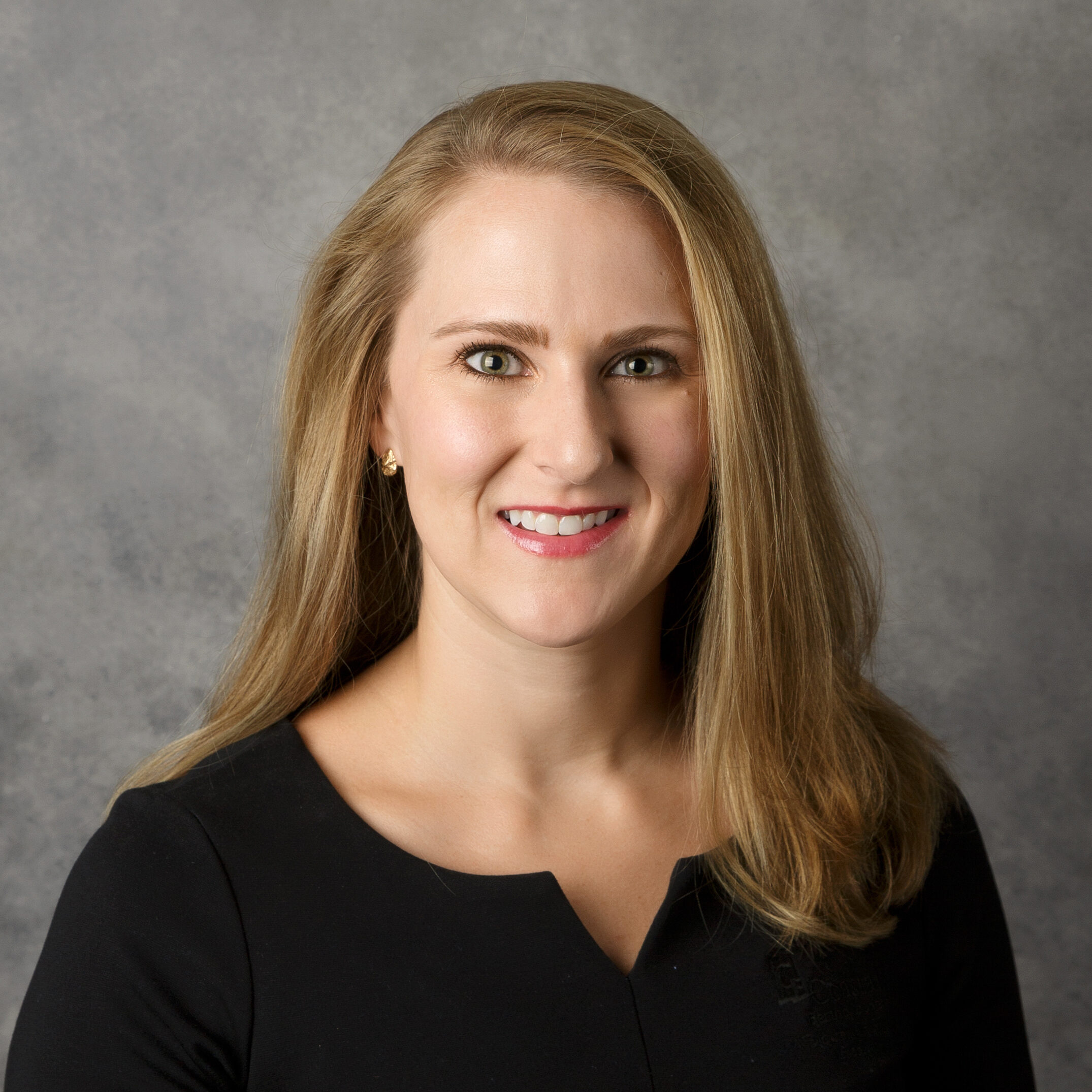 The image shows a person with long hair, smiling, wearing a black top against a neutral background.