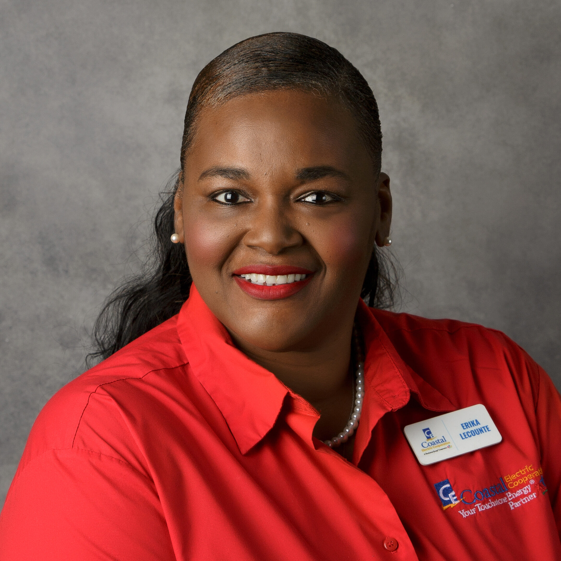 A person smiling in a red shirt with a name tag, set against a plain gray background. No landmarks or historical buildings visible.