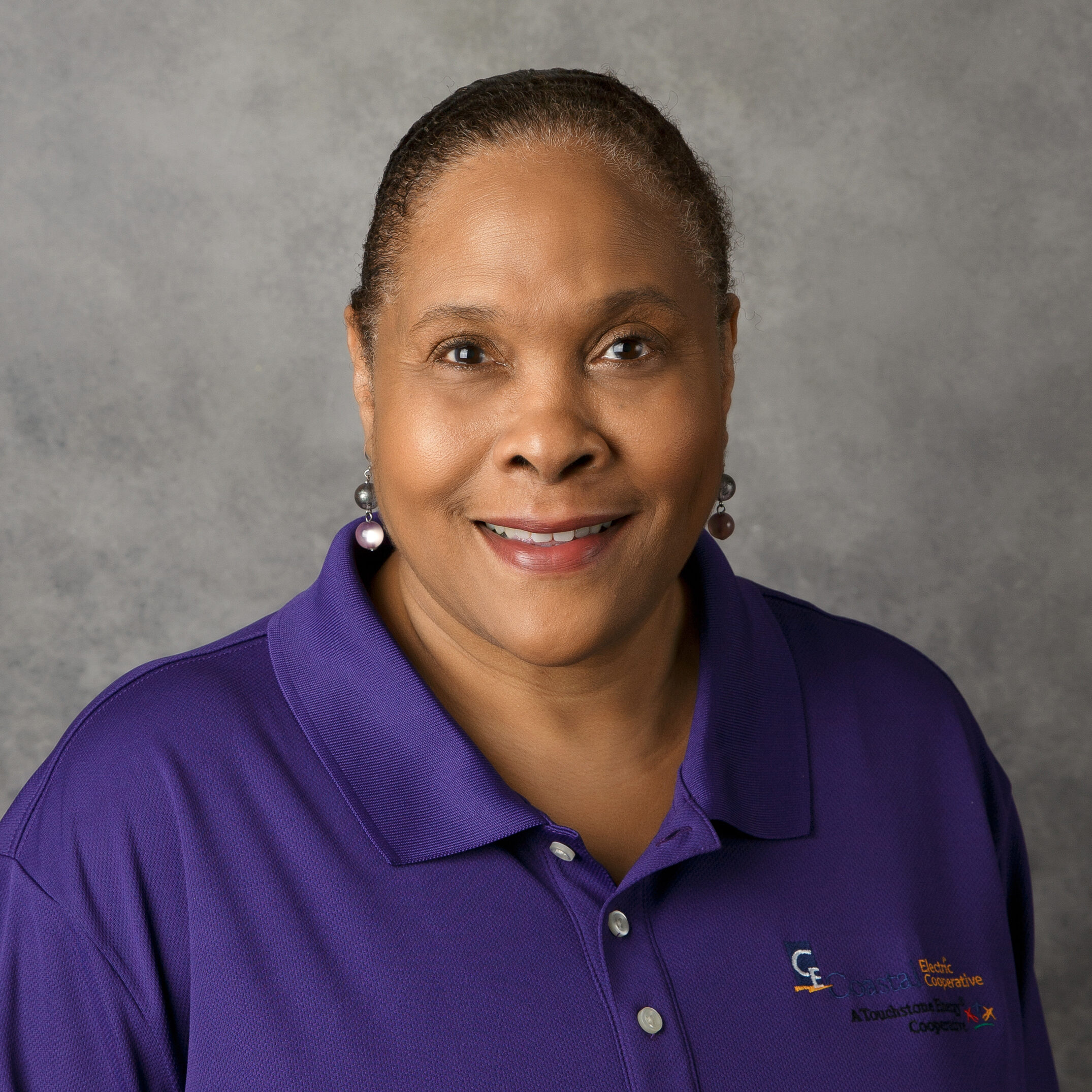 A person in a purple shirt, smiling against a plain gray background. Shirt has a logo and text on the left side.