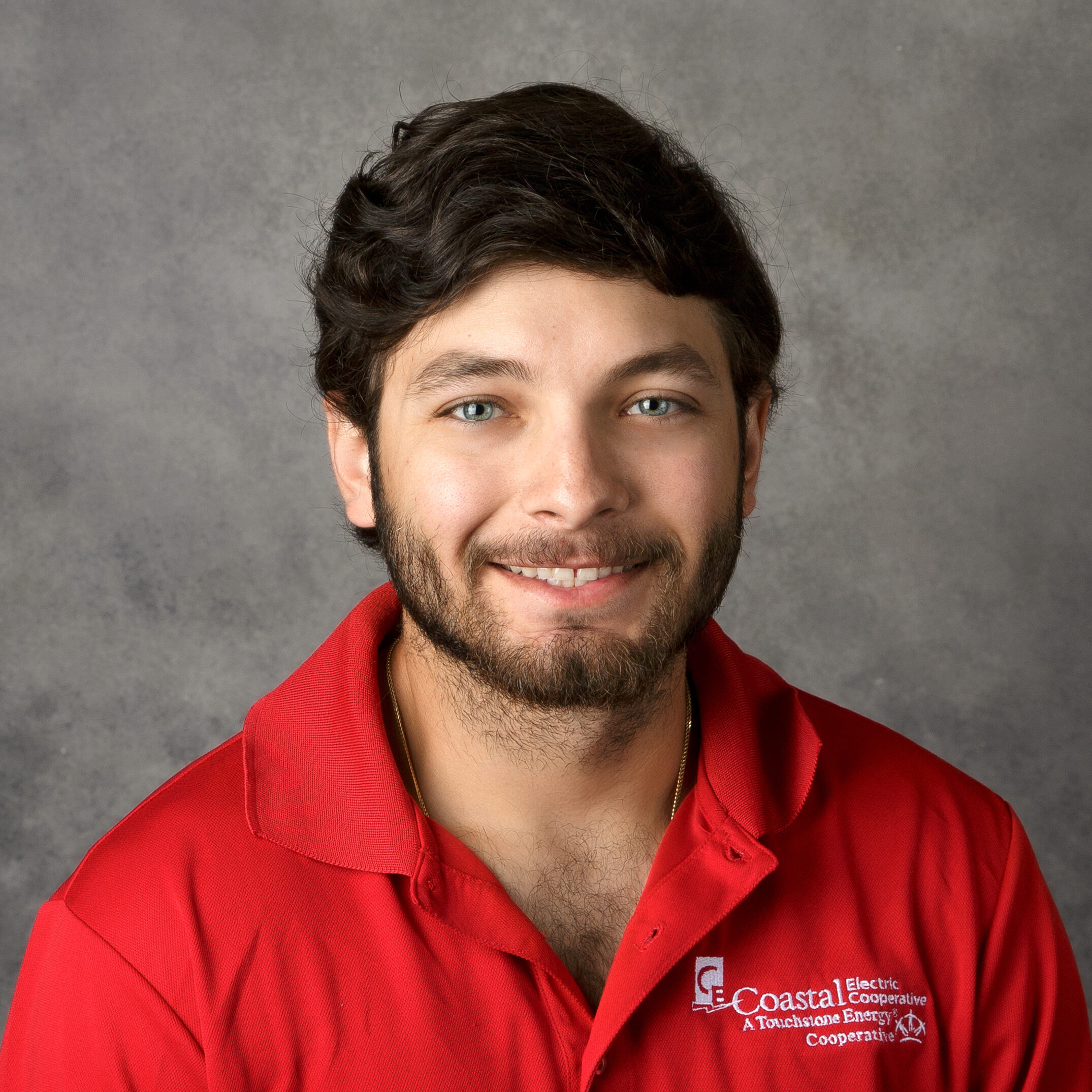 A person with a beard smiles in front of a gray background, wearing a red polo with a logo reading "Coastal Electric Cooperative."