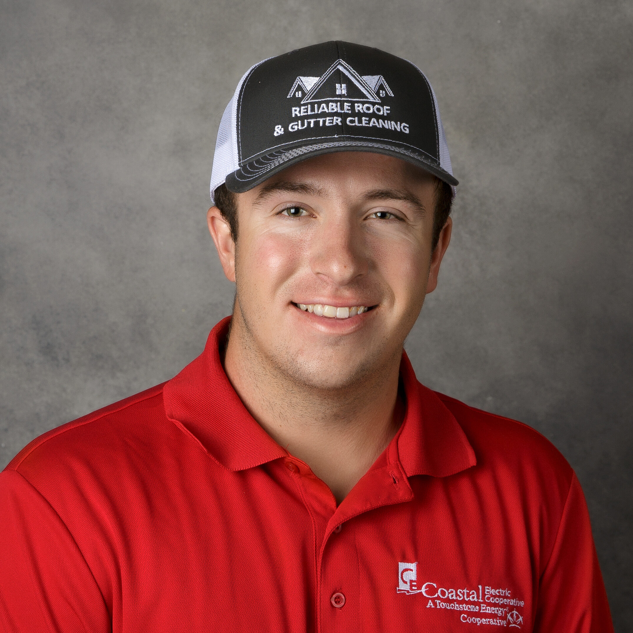 A person in a red shirt and cap with "RELIABLE ROOF & GUTTER CLEANING" branding, smiles against a plain gray background.