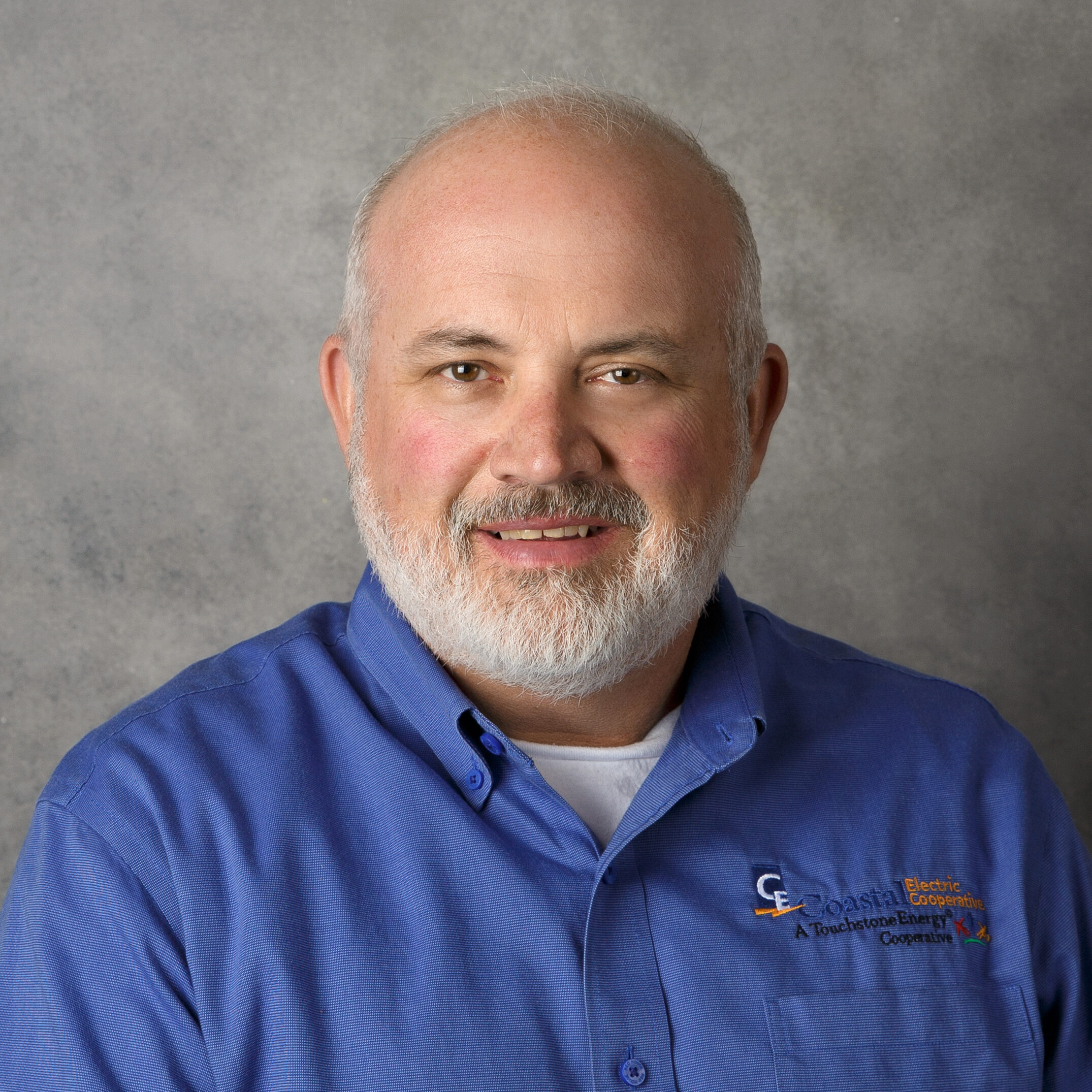 A person with a beard and blue shirt is smiling against a gray backdrop. The shirt has an embroidered logo on it.