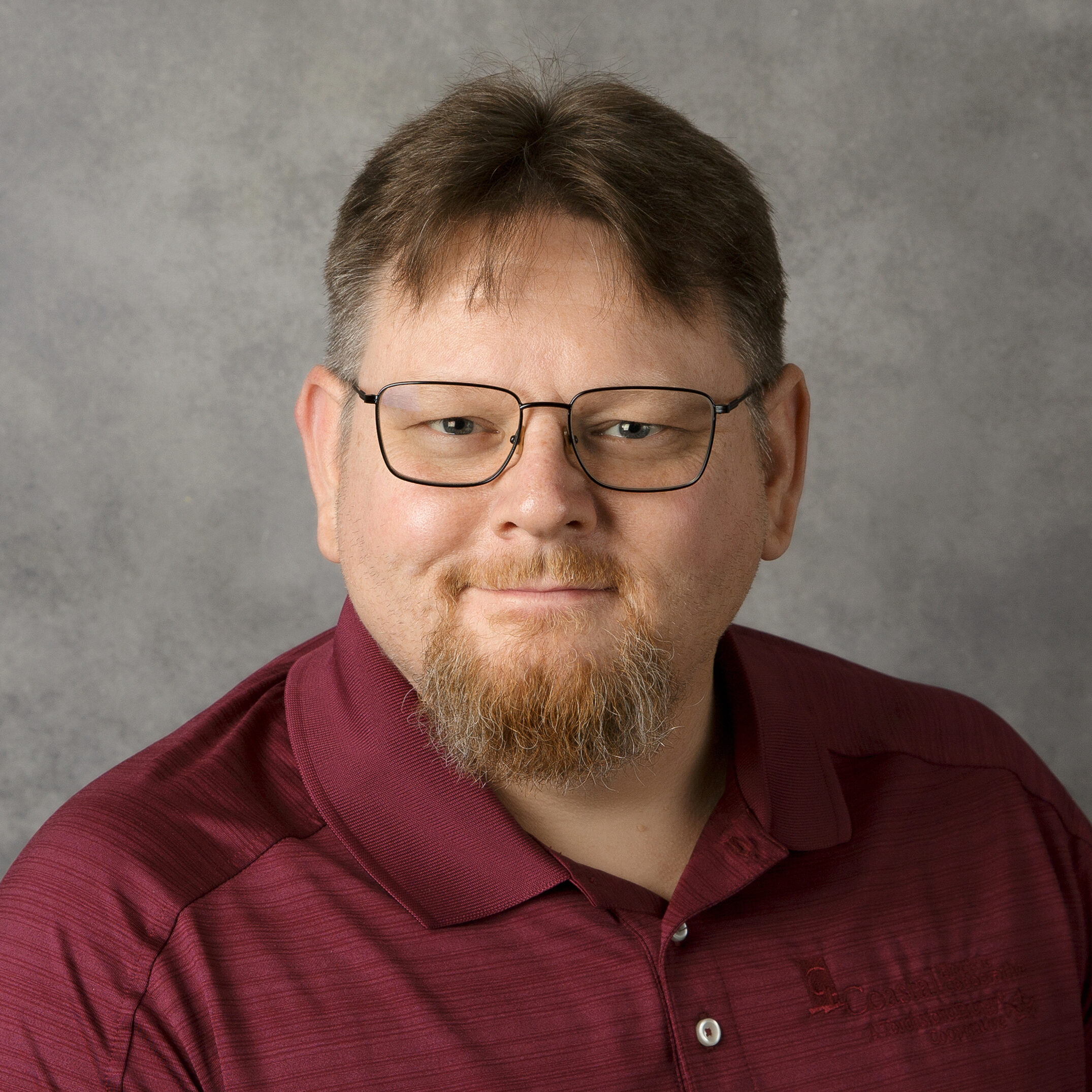 A person with glasses and facial hair wears a maroon collared shirt, posing against a gray background, smiling gently at the camera.