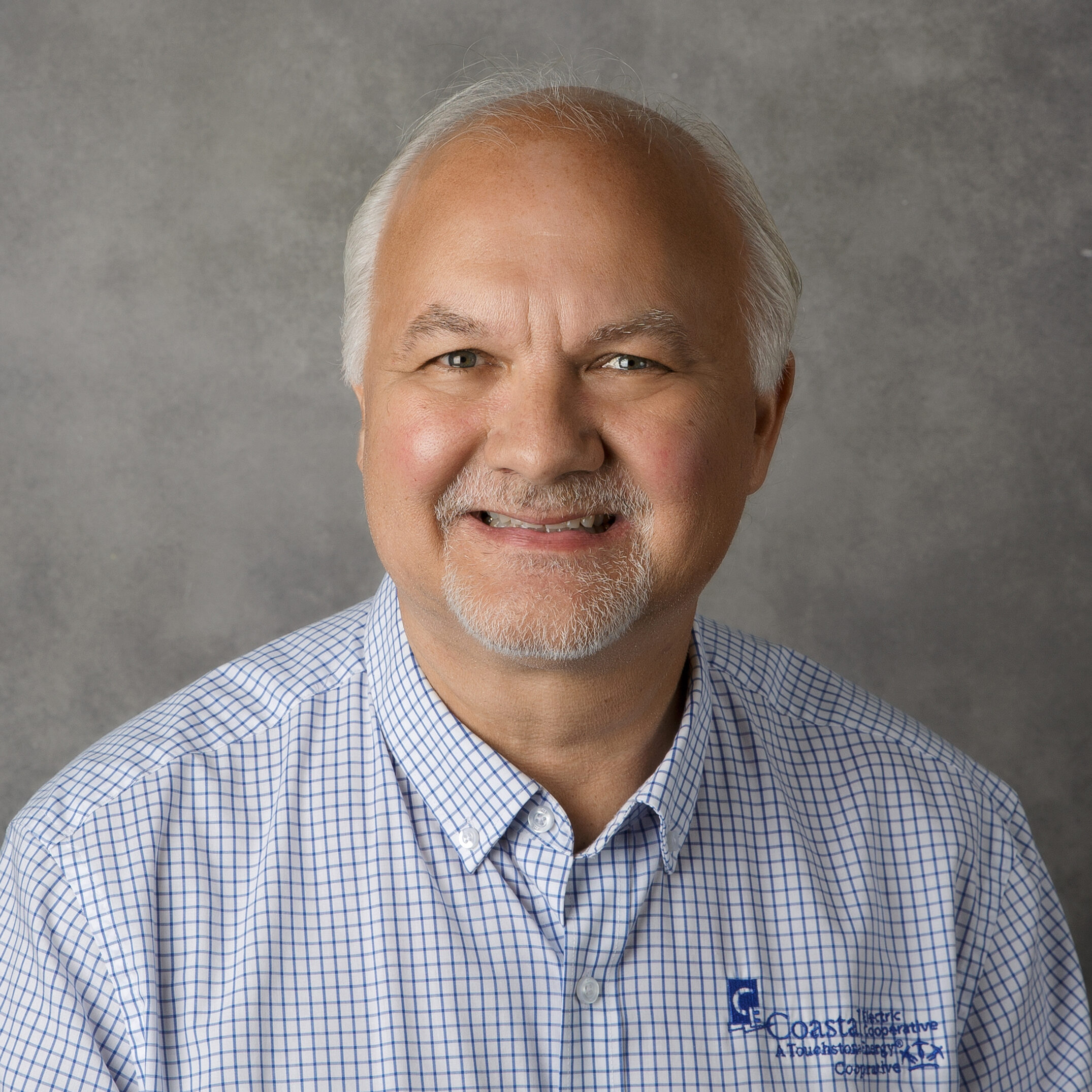 A person with gray hair and a beard smiles, wearing a checked shirt with a company logo, set against a gray background.