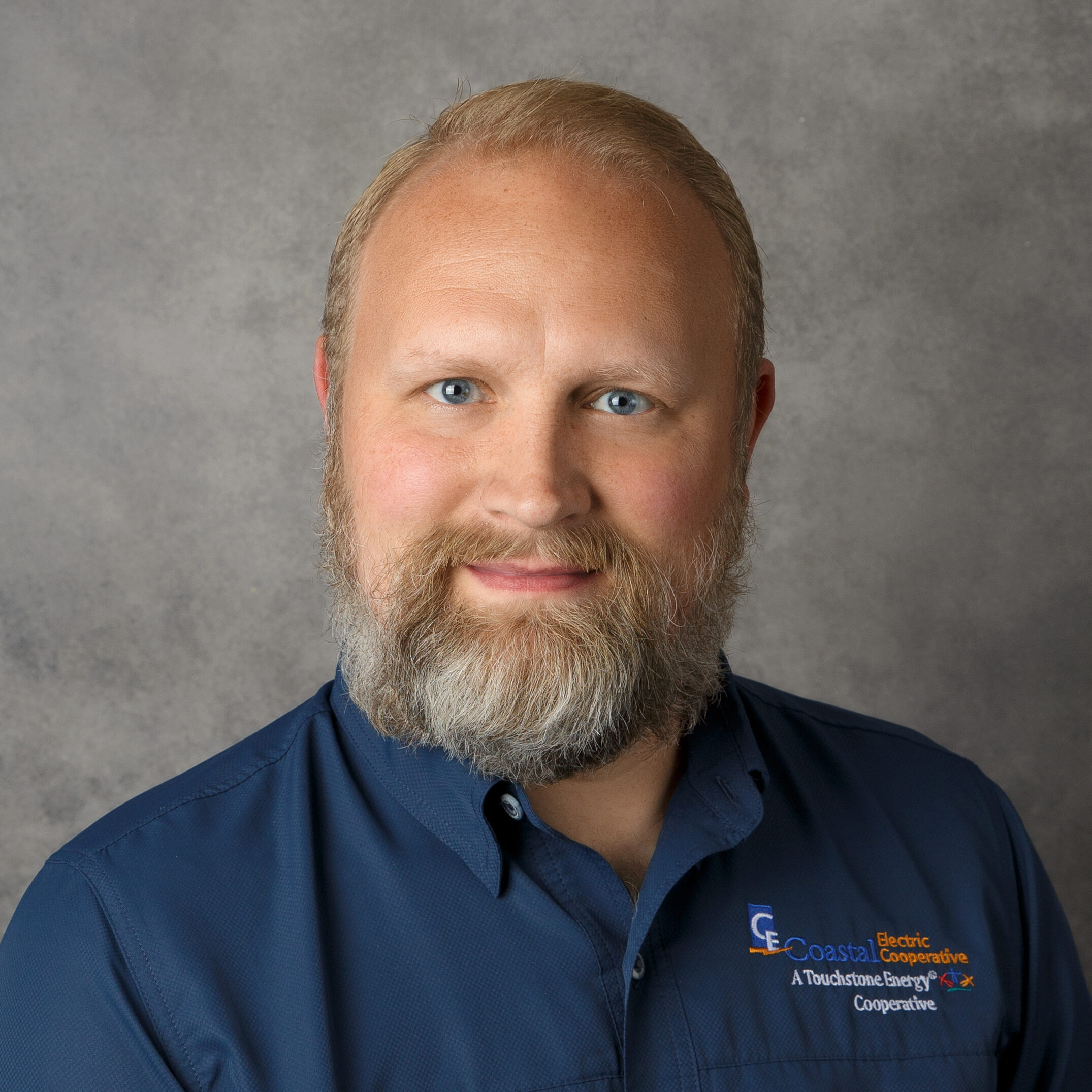 A person with a beard wearing a blue shirt with "Coastal Electric Cooperative" logo. Neutral gray background. Facial expression is calm and friendly.