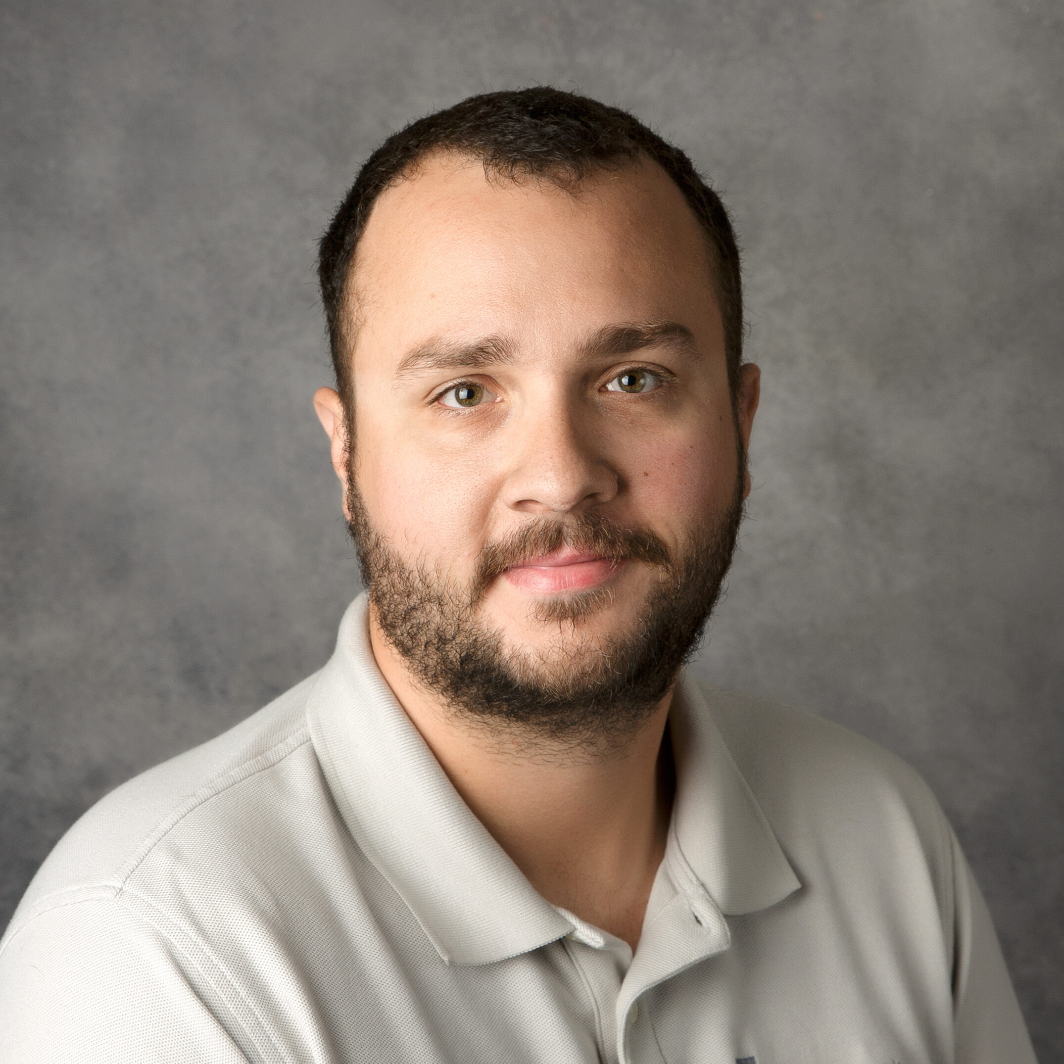 A person with short hair and a beard wearing a light-colored polo shirt, posing against a plain gray background.