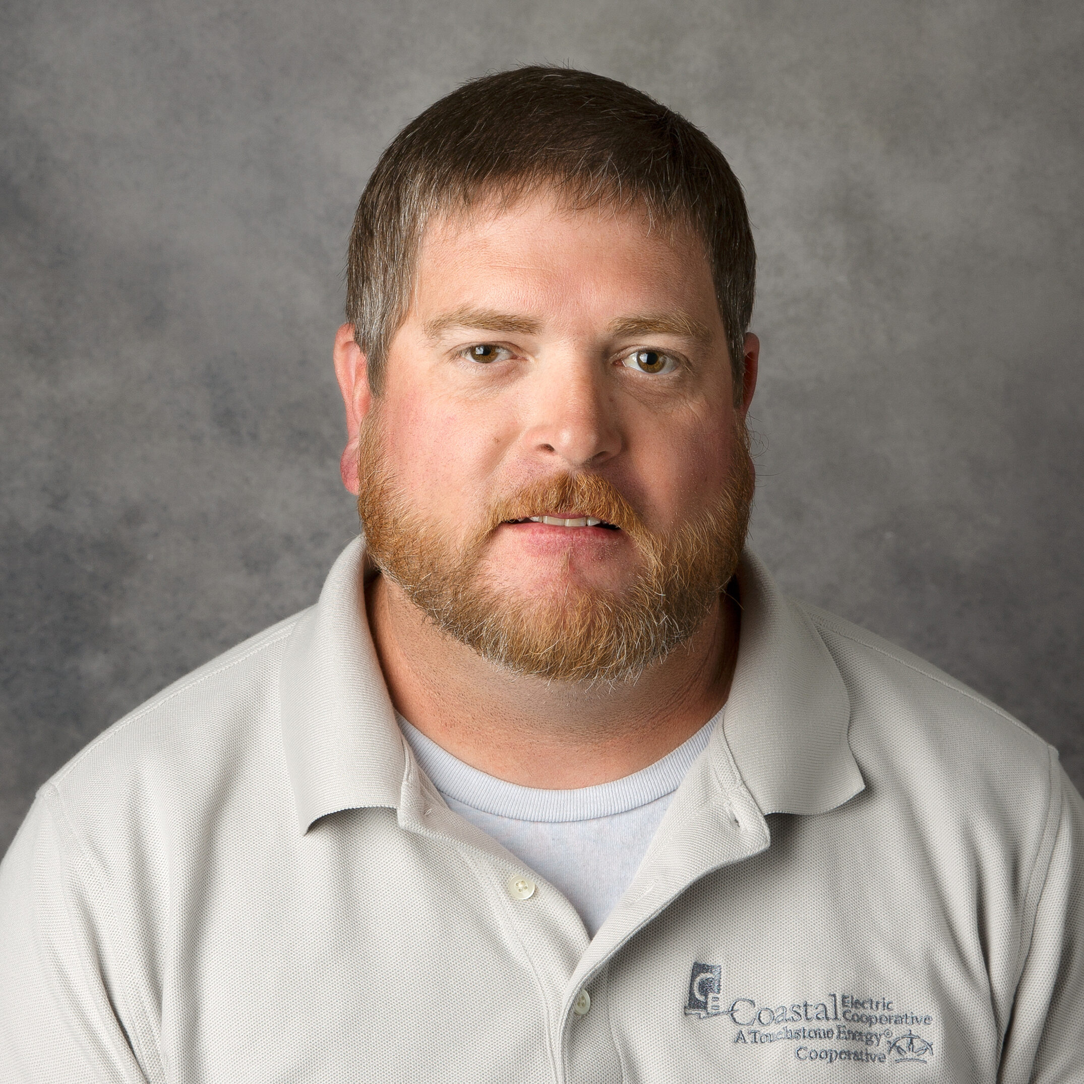 A person with a beard wears a grey polo shirt with "Coastal Electric Cooperative" logo, against a neutral gray background.