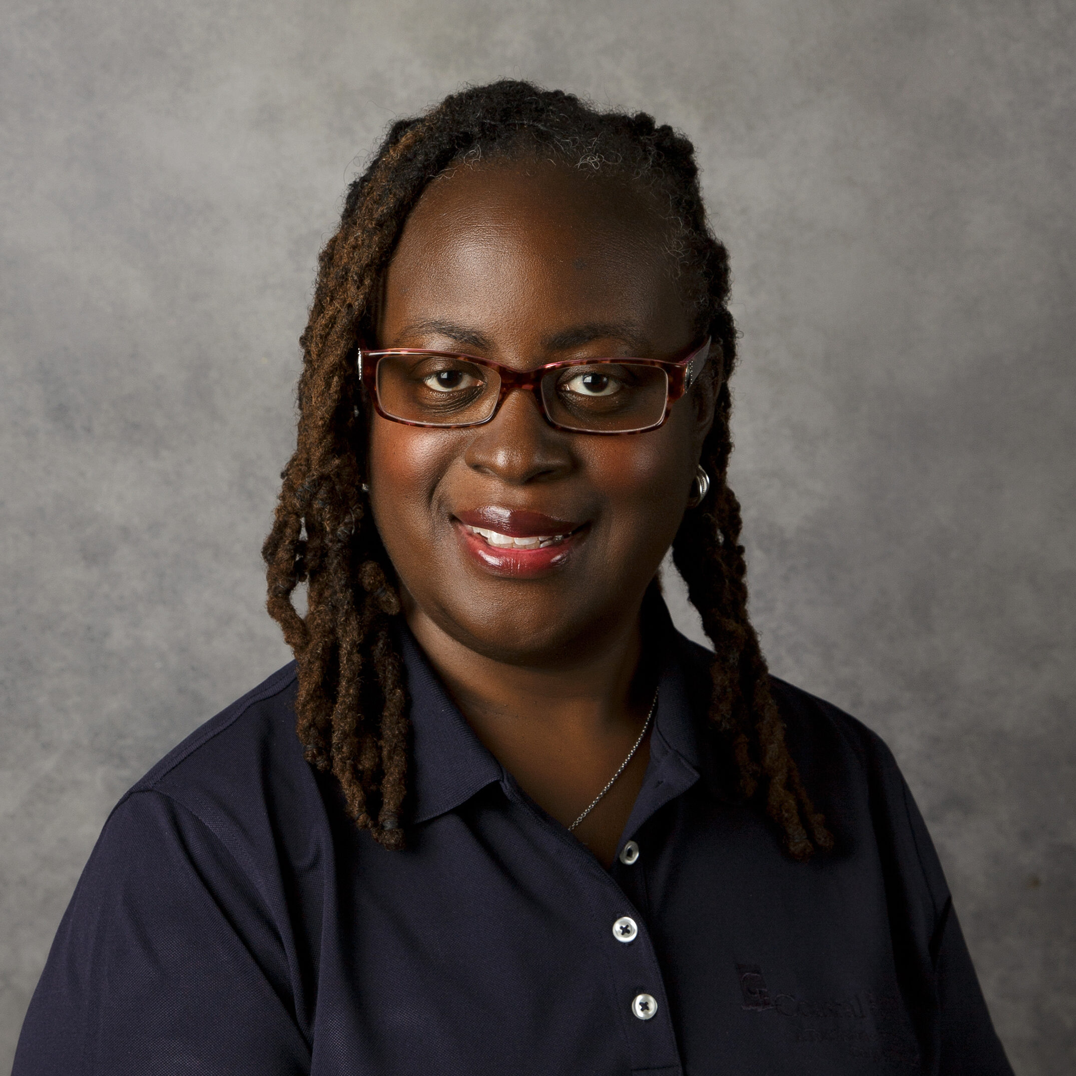 A person with glasses, wearing a dark shirt, smiles in front of a neutral gray background. Hair is styled in long twists.