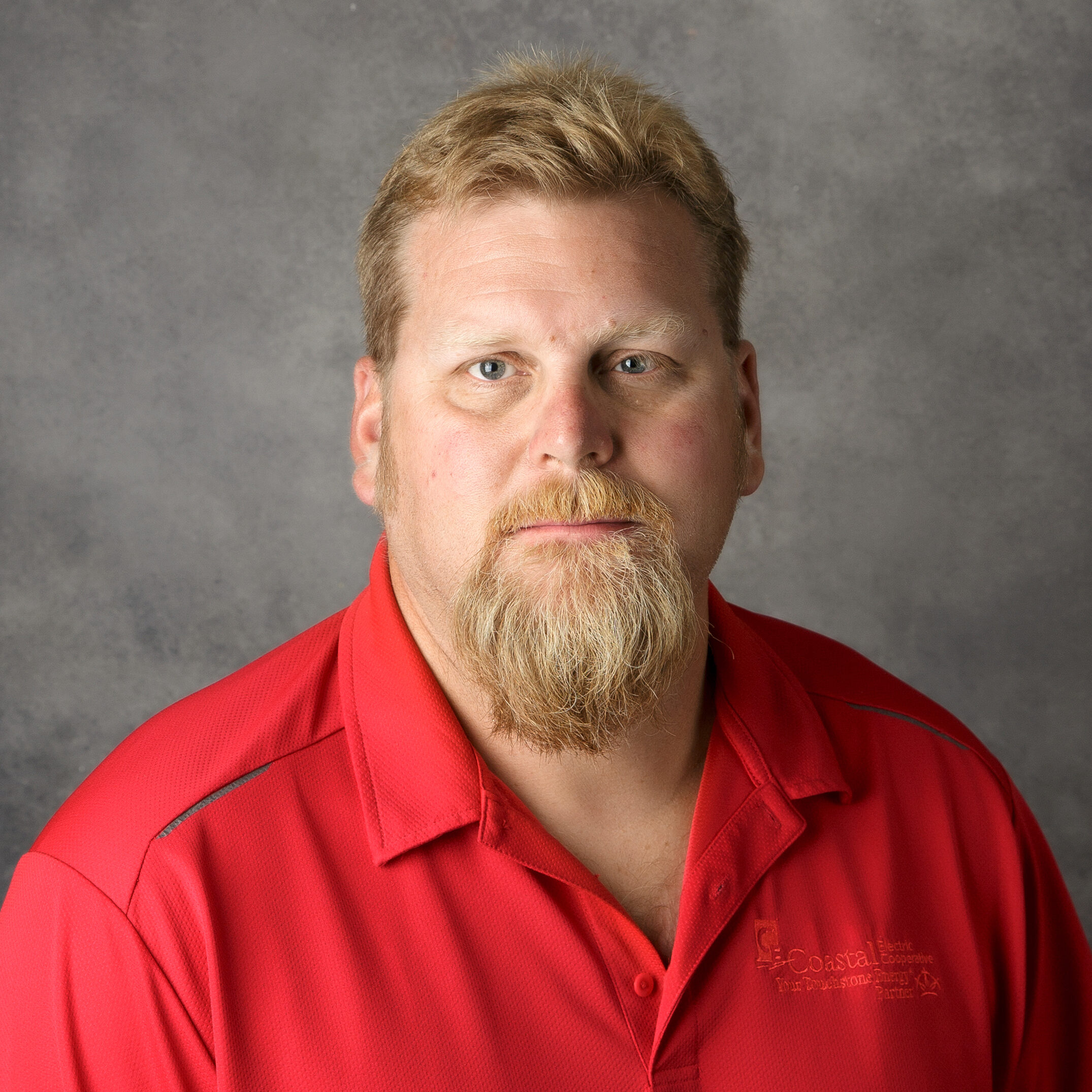 A person with a light beard and short hair is wearing a red shirt. The background is a plain, gray surface.