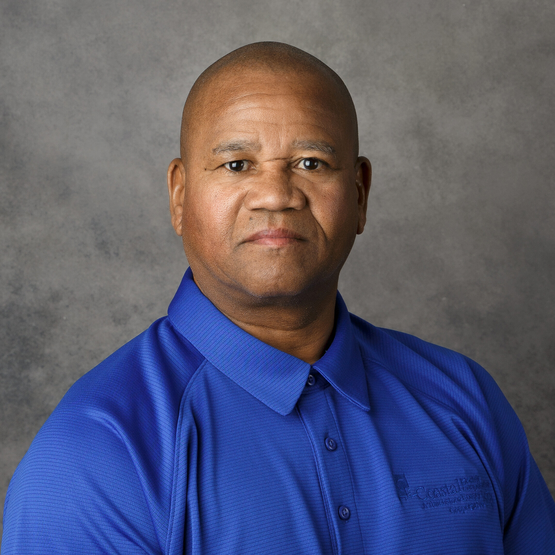 A person in a blue polo shirt poses against a gray background, looking directly at the camera. No landmarks or historical buildings present.