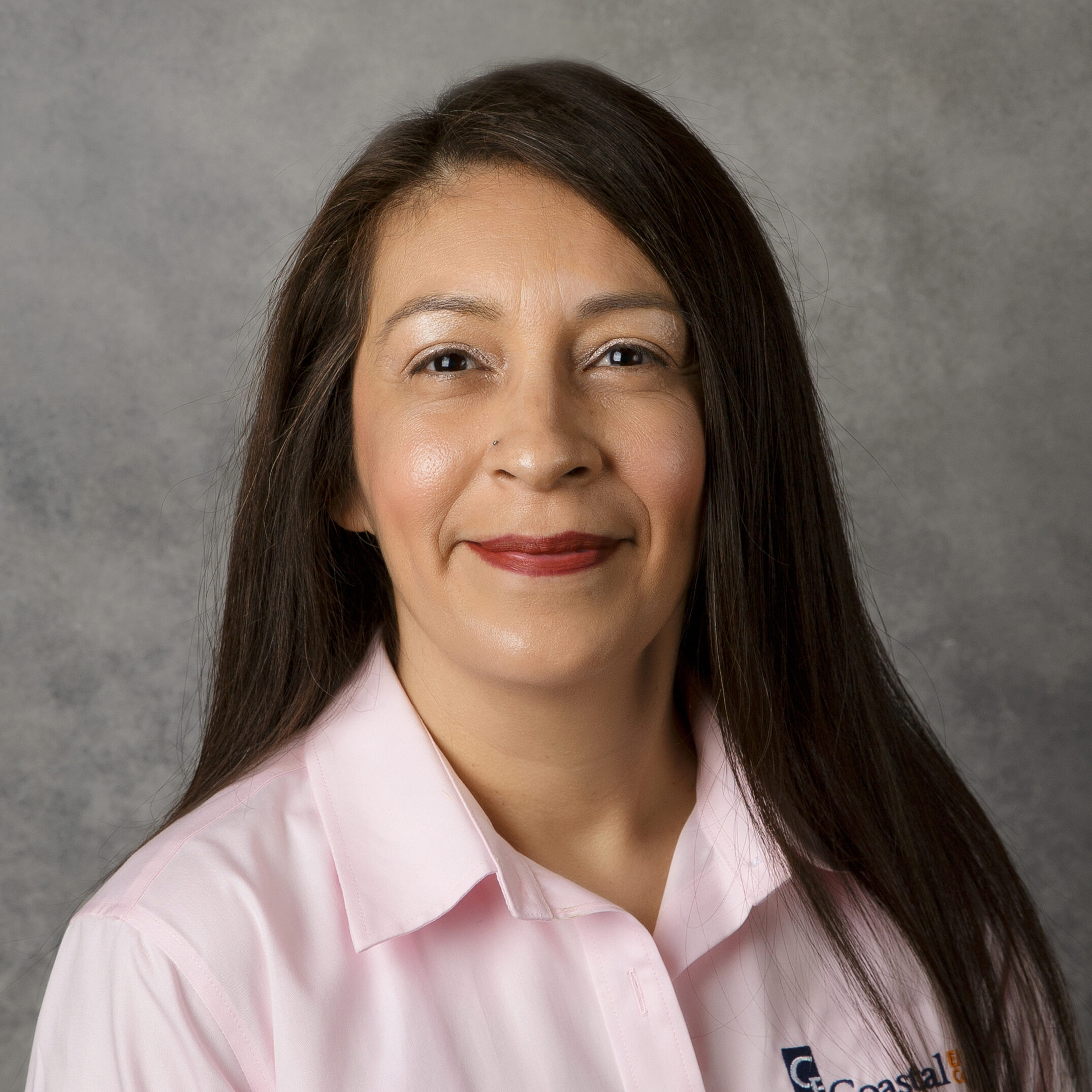 A person with long hair, wearing a pink shirt with insignia, smiles against a gray background. No landmarks or historical buildings present.