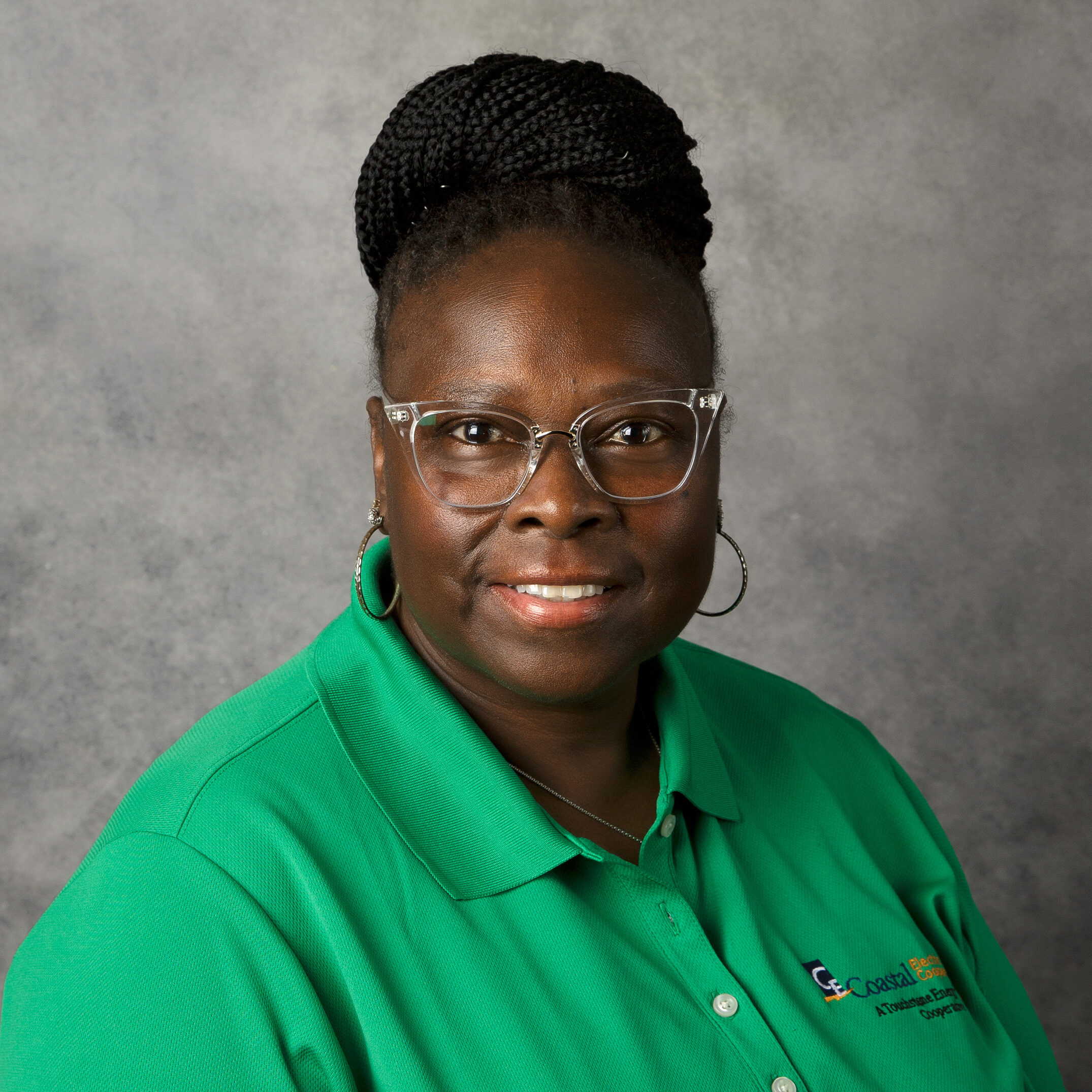 A person with braided hair and glasses smiles, wearing a green polo shirt with a logo. Gray background.