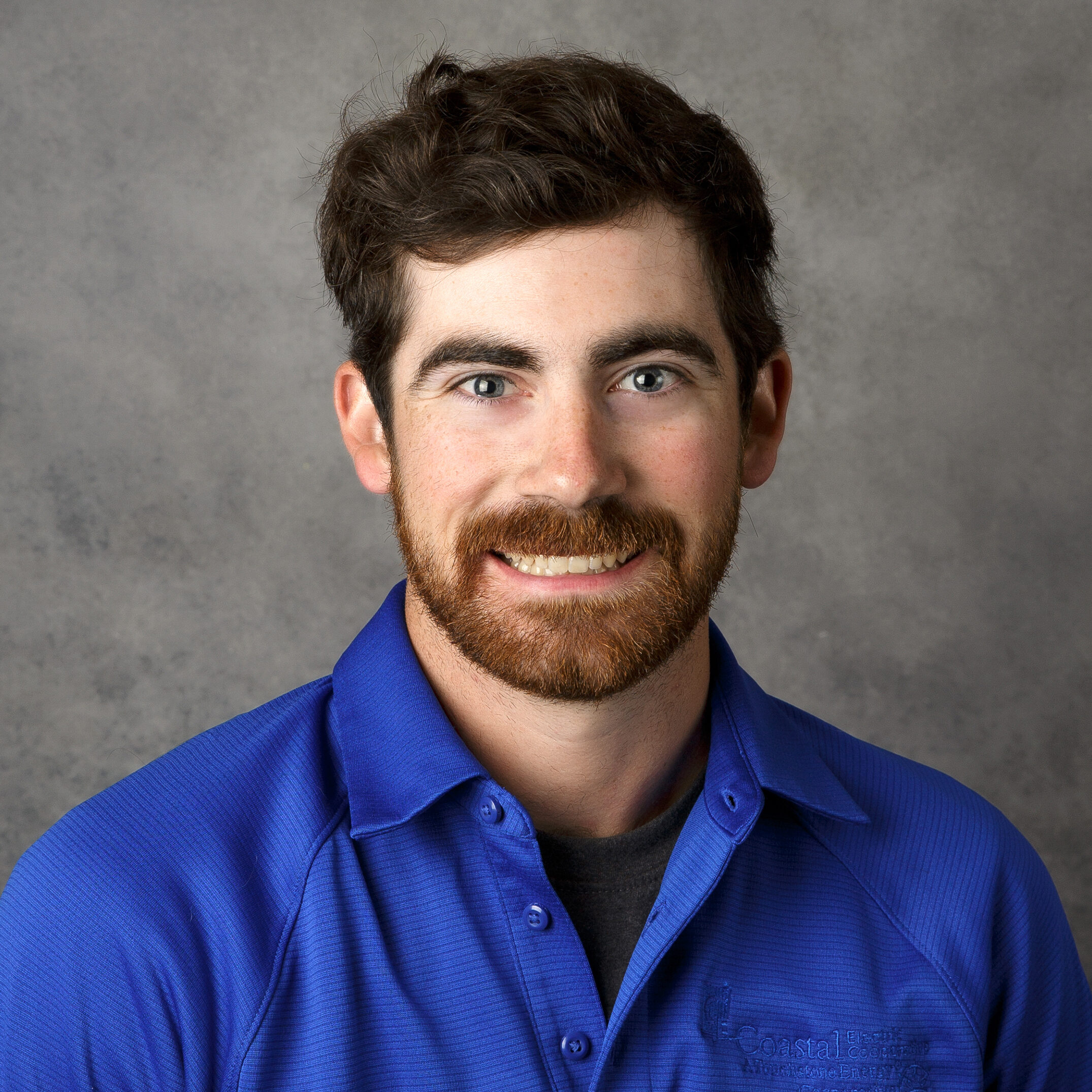 A person with a beard smiles, wearing a blue shirt with a logo. Grey background. No visible landmarks or historical buildings.