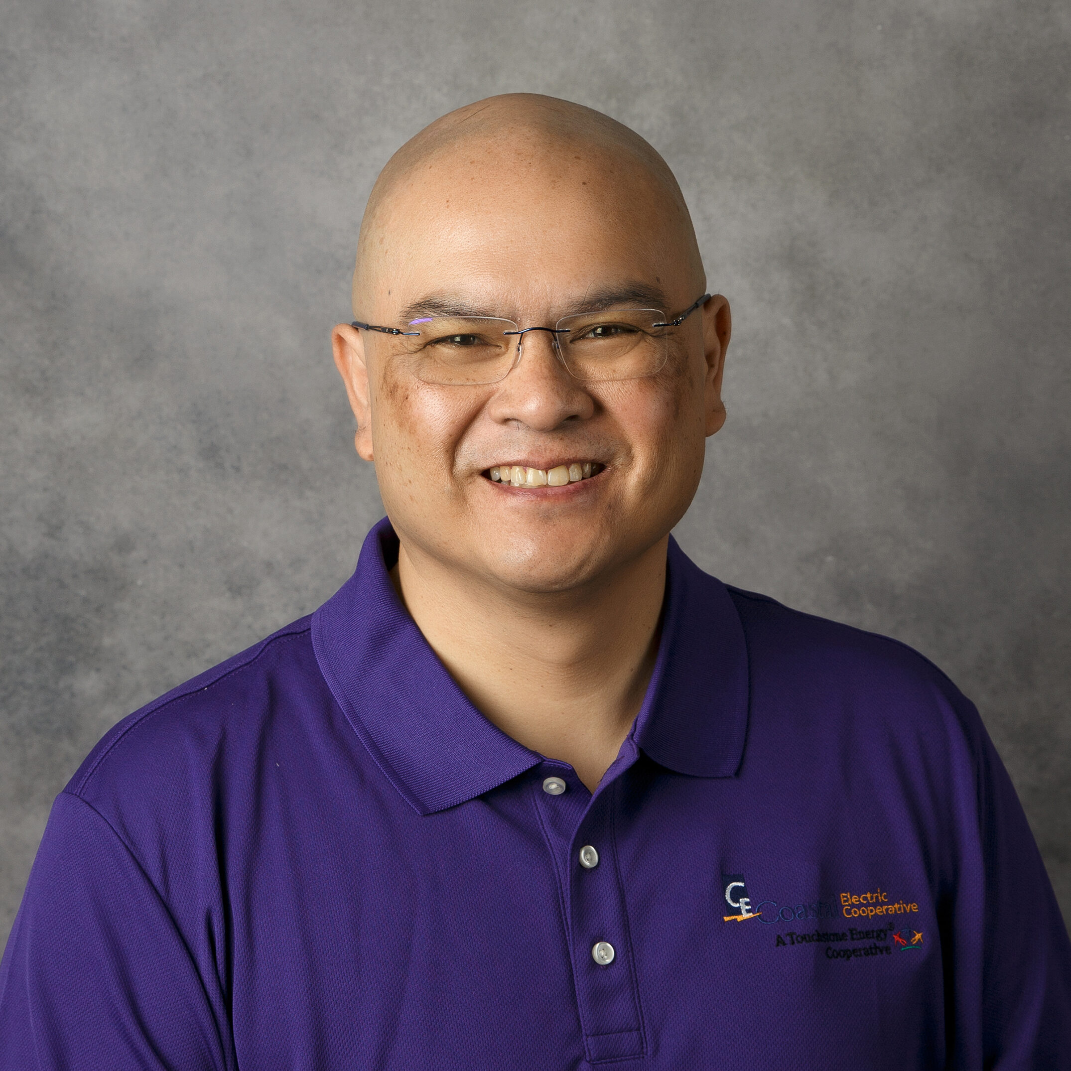 A smiling person with glasses and a purple polo shirt stands in front of a gray textured background.