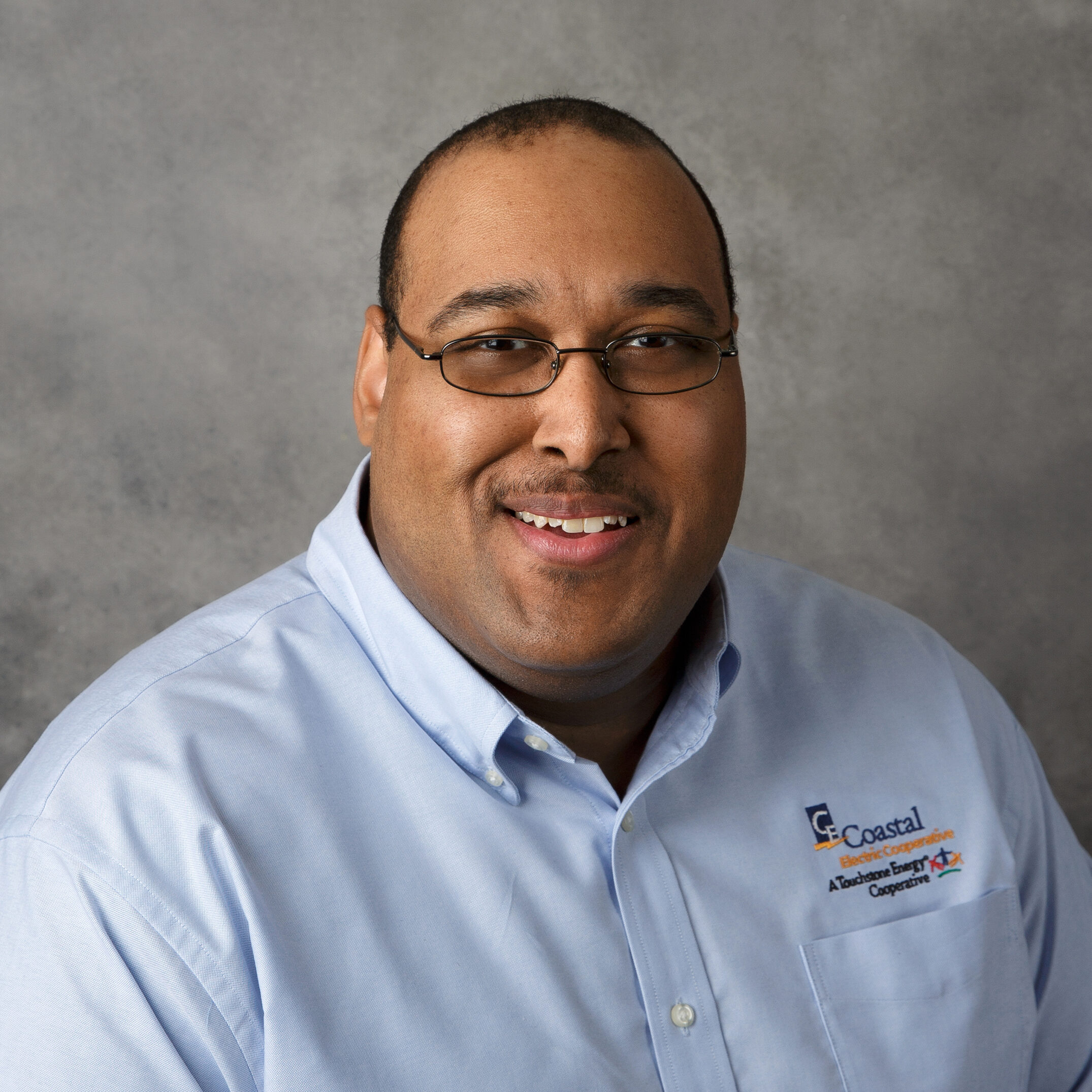 A person with glasses smiles, wearing a light blue shirt featuring a logo, against a plain gray background. No landmarks or buildings visible.