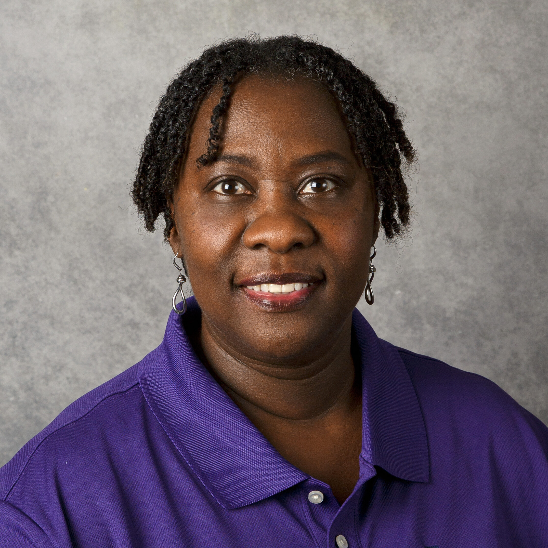 A person wearing a purple shirt smiles in front of a gray background, displaying short curly hair and earrings.