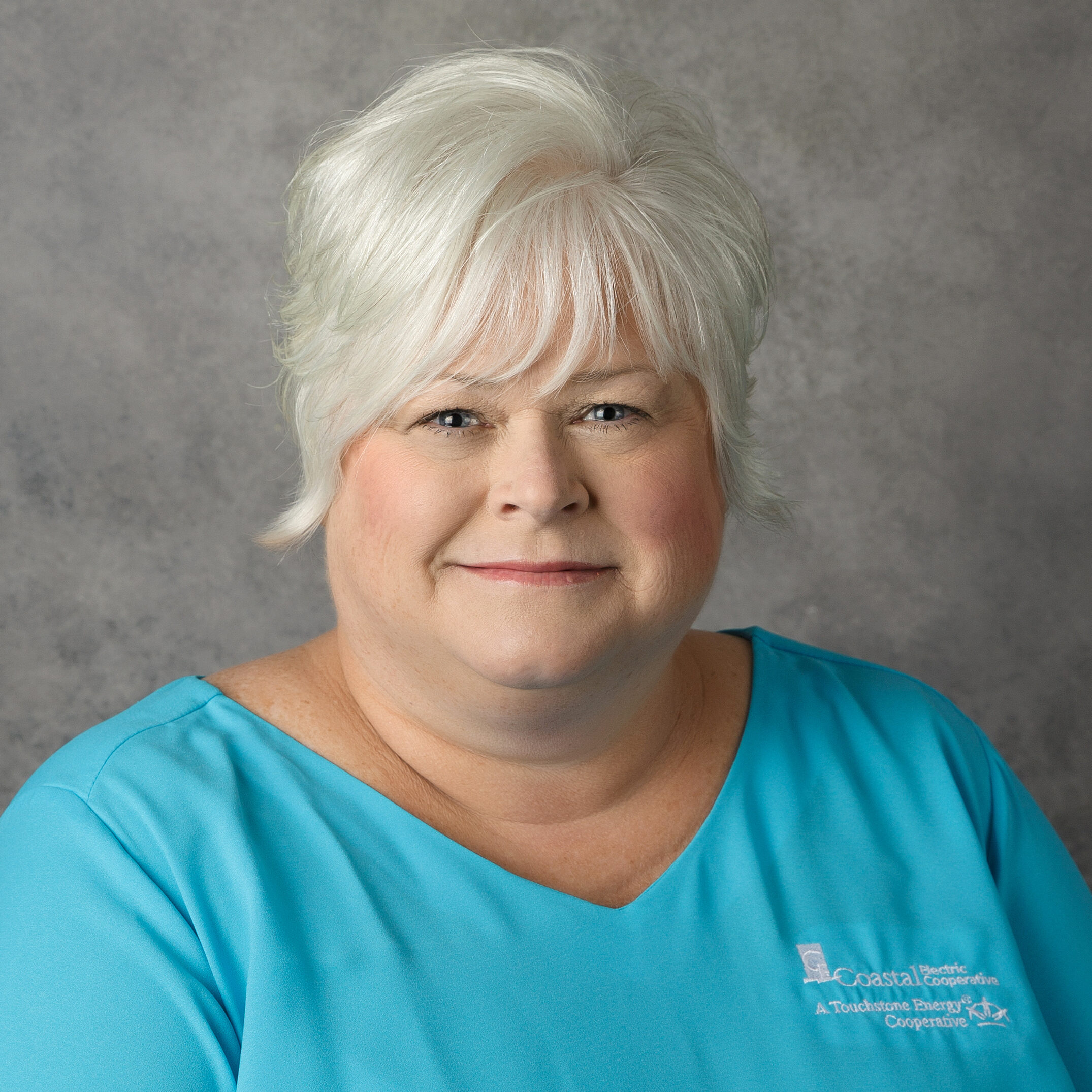 A person with short, white hair is wearing a blue shirt with a logo, posed against a neutral gray background.