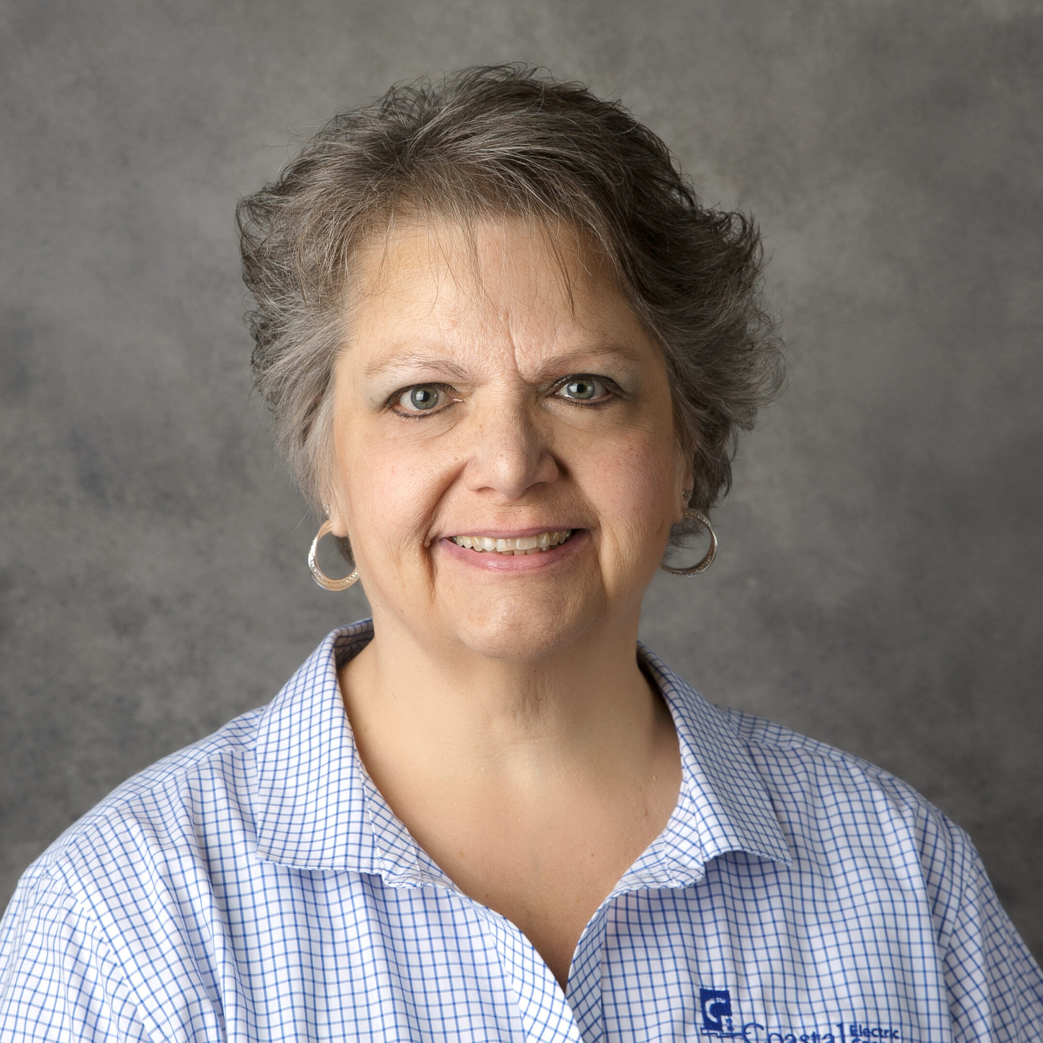 A person with short gray hair smiles, wearing a blue plaid shirt with a logo, against a gray backdrop.