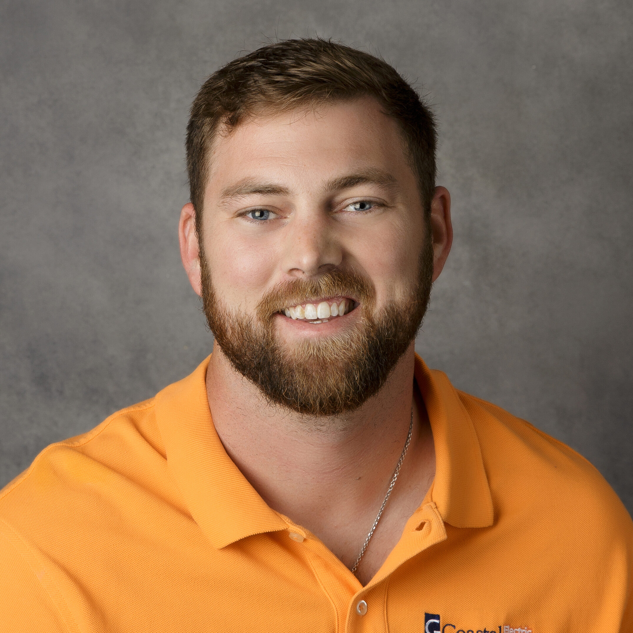 A person with a beard smiles at the camera, wearing an orange collared shirt. The background is plain gray.