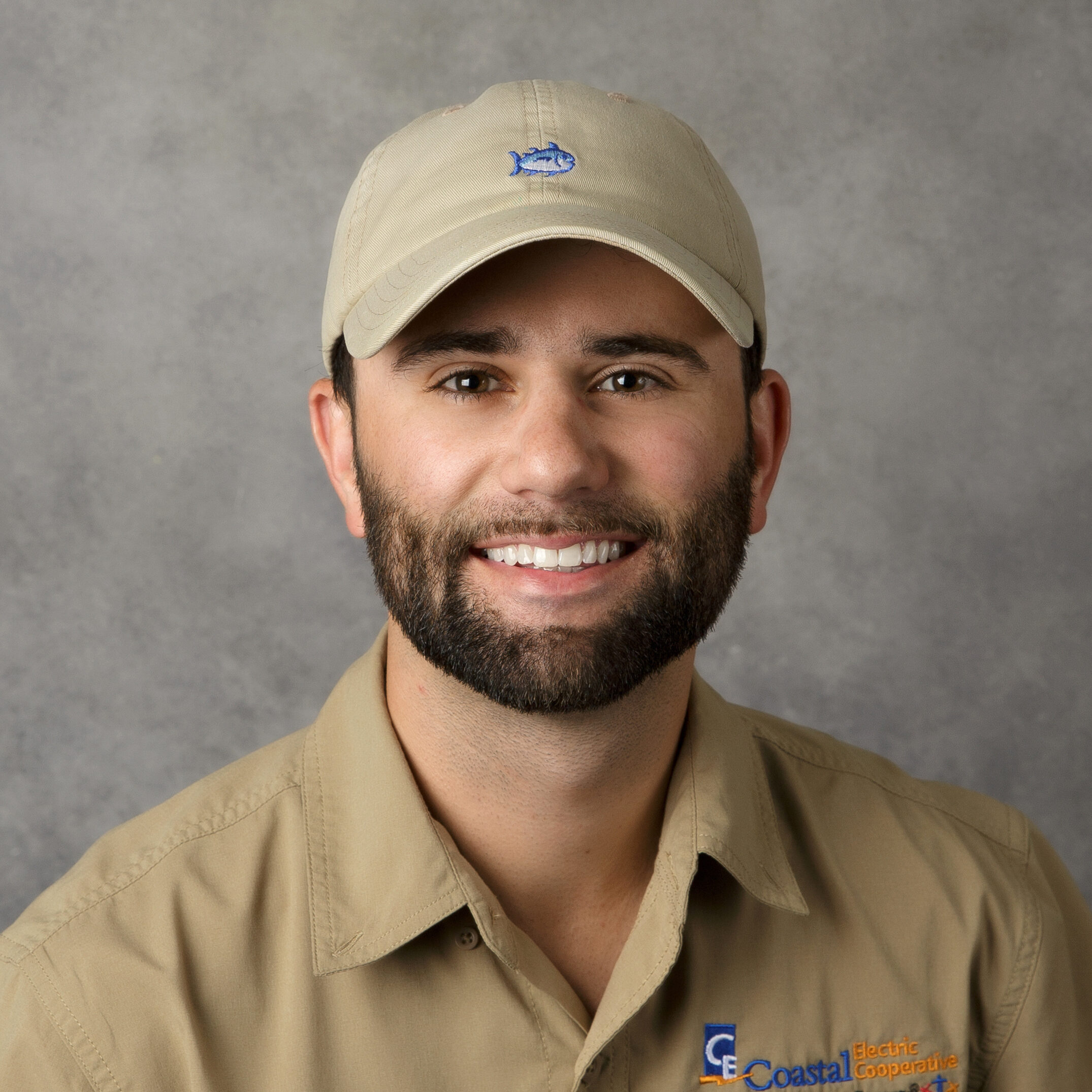 A person in a beige shirt and cap smiles against a gray background. The shirt has an embroidered logo on the left side.