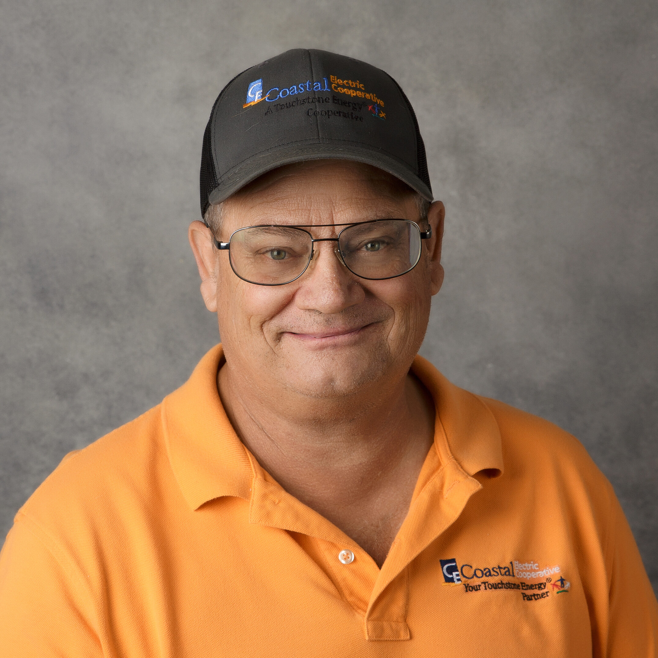 Person wearing glasses, a black cap, and an orange polo with "Coastal Electric Cooperative" logo against a plain gray background. Smiling expression.