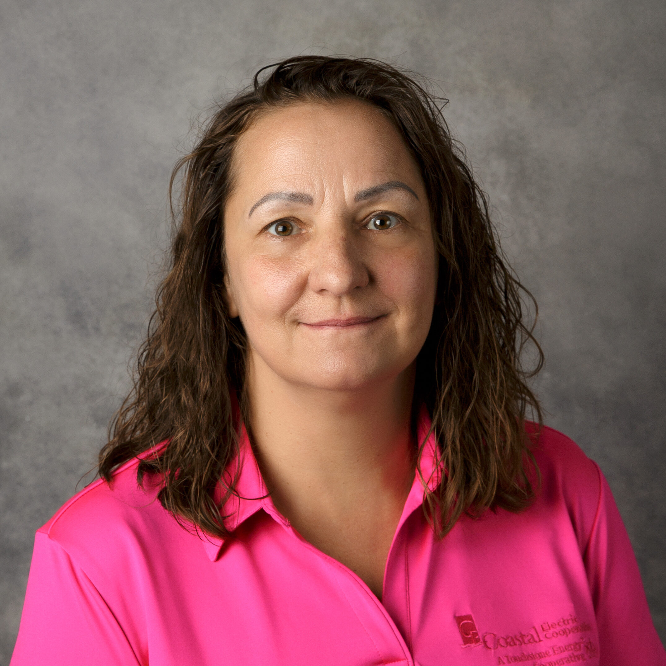 A person with curly hair, wearing a pink shirt, stands against a gray background. No landmarks or historical buildings are visible.