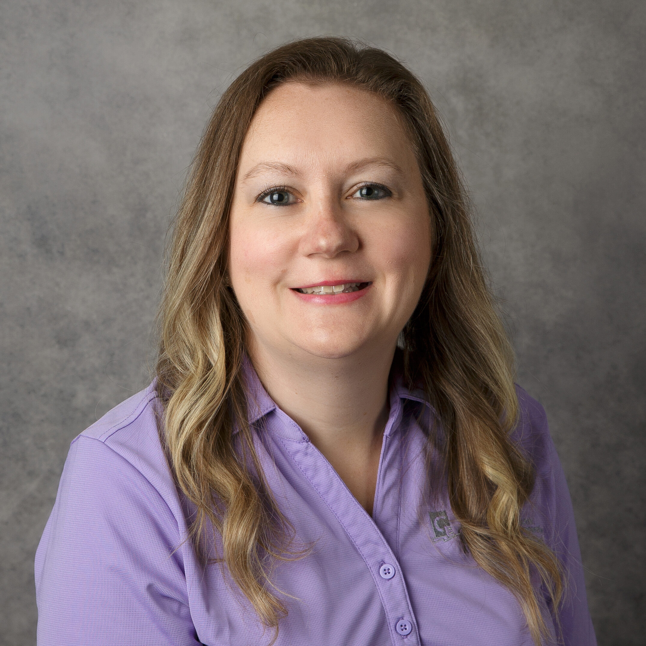 A person with long hair wearing a purple shirt smiles against a plain gray background, creating a simple and professional portrait.