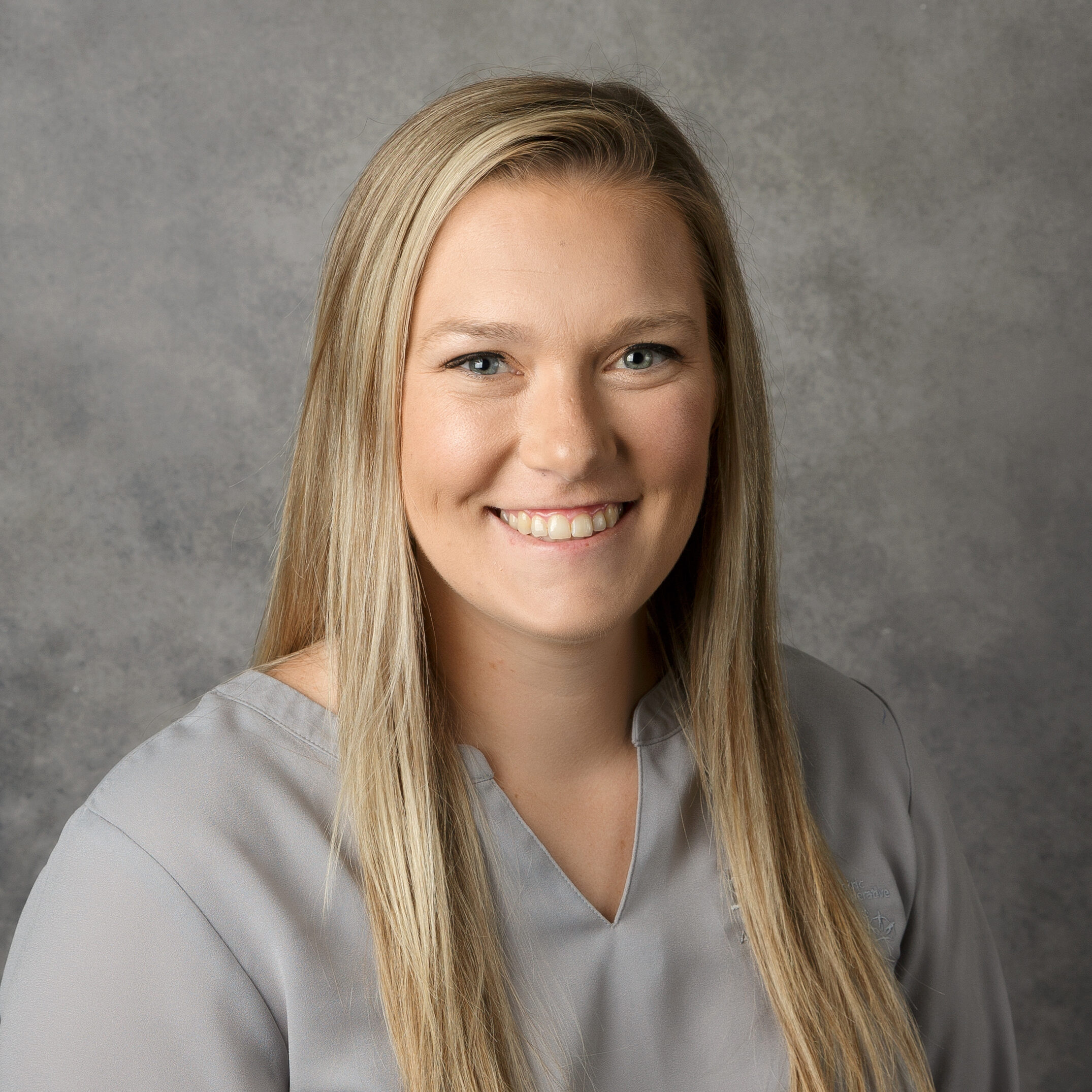 A person with long hair smiles against a neutral gray background, wearing a light gray shirt. The image is a portrait style photograph.