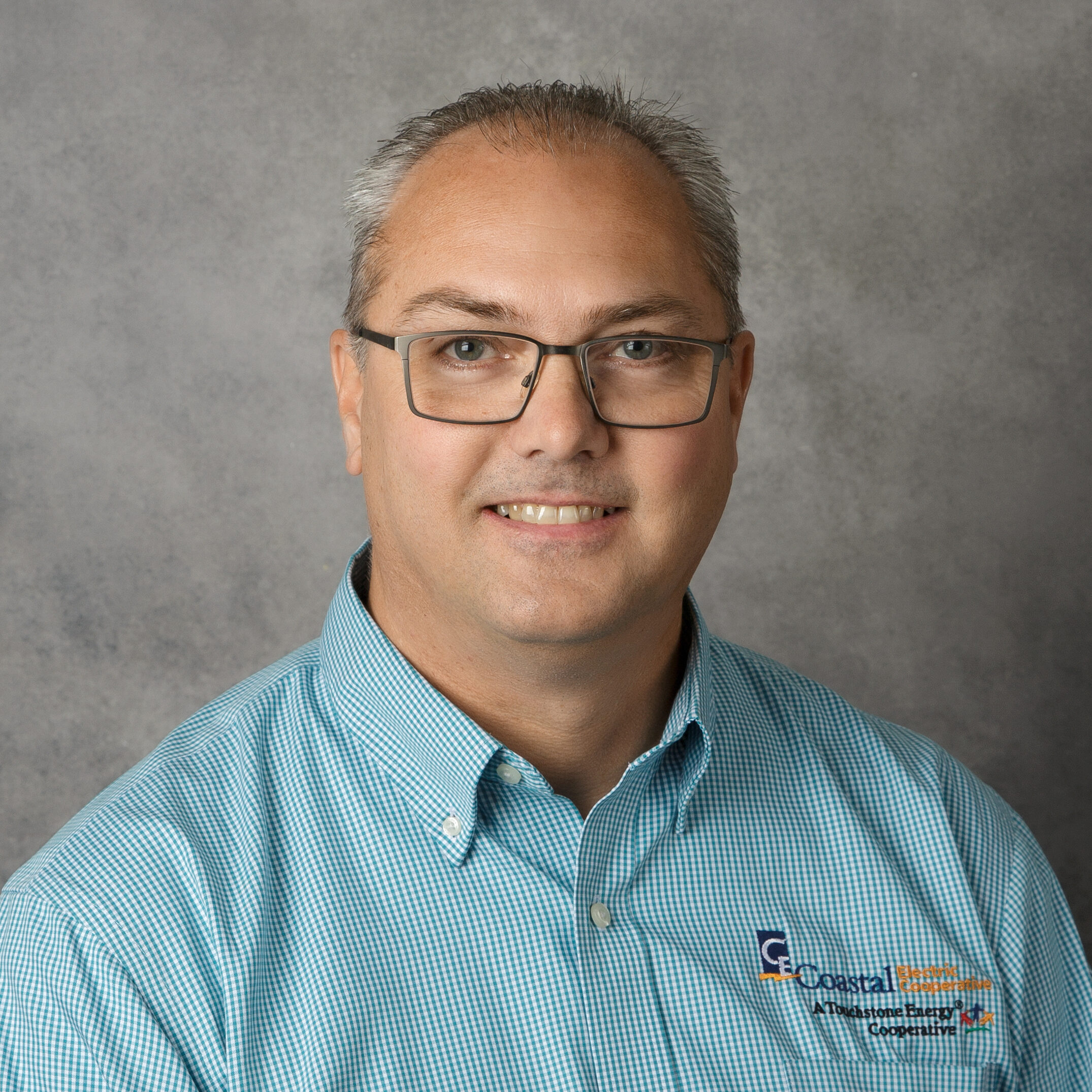 A person with glasses and short hair wearing a blue collared shirt with a logo, set against a gray background.