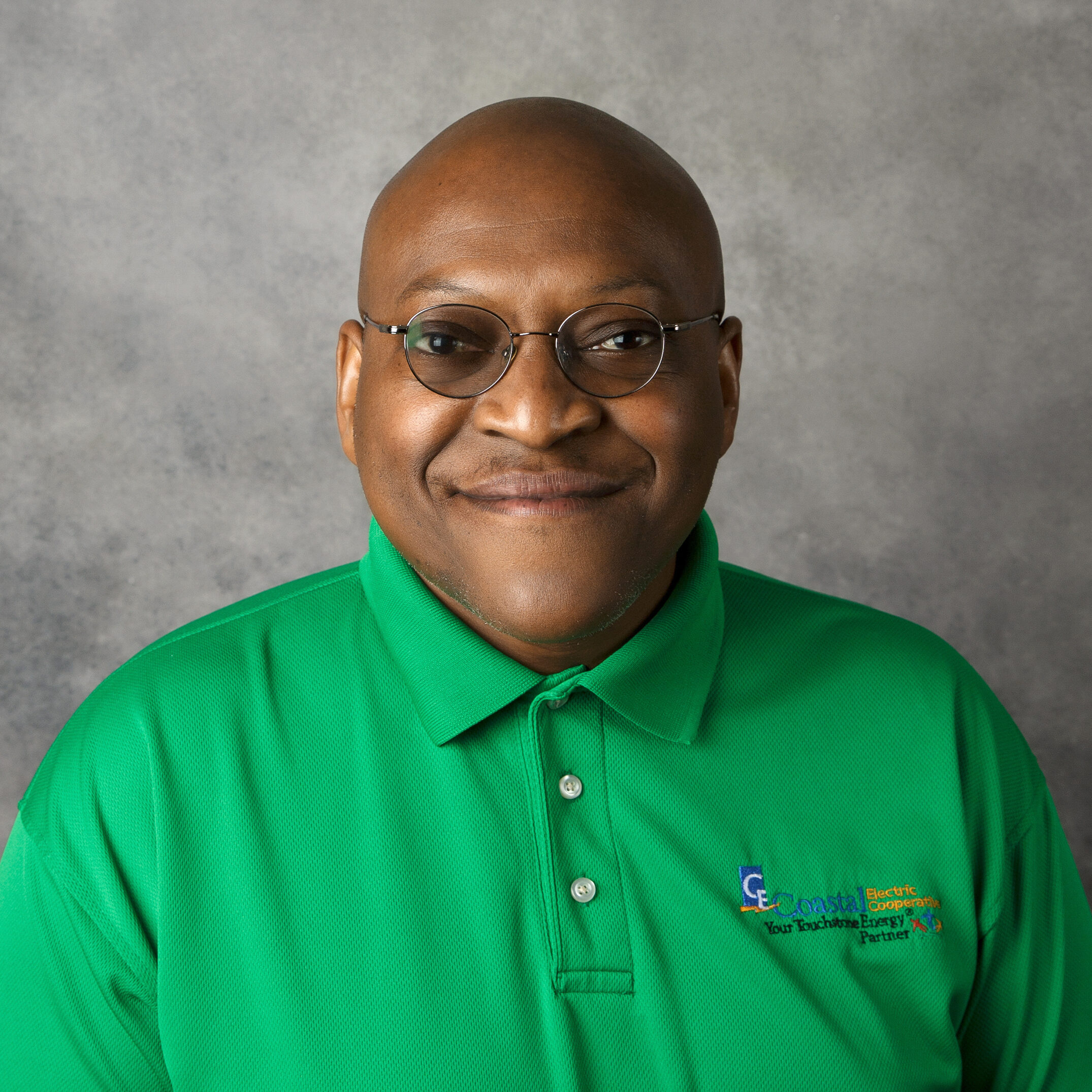 A person wearing glasses and a green polo shirt, standing against a plain gray background, is smiling at the camera.