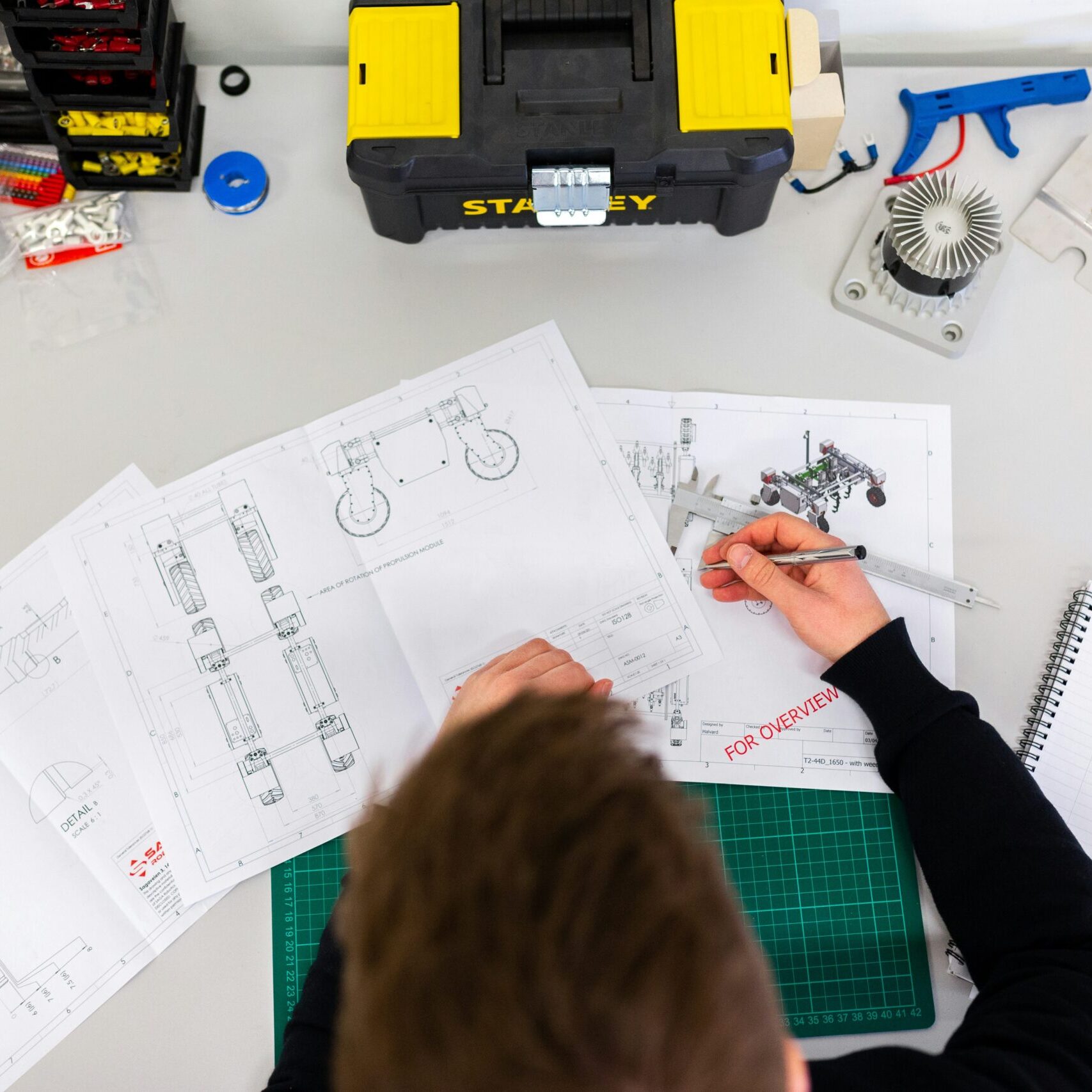 A person is working at a desk with technical drawings and tools, including a caliper, toolbox, and notebook.