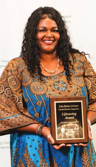 A person in patterned clothing holds a wooden award plaque. Background features a repeating green and white text logo on a light backdrop.